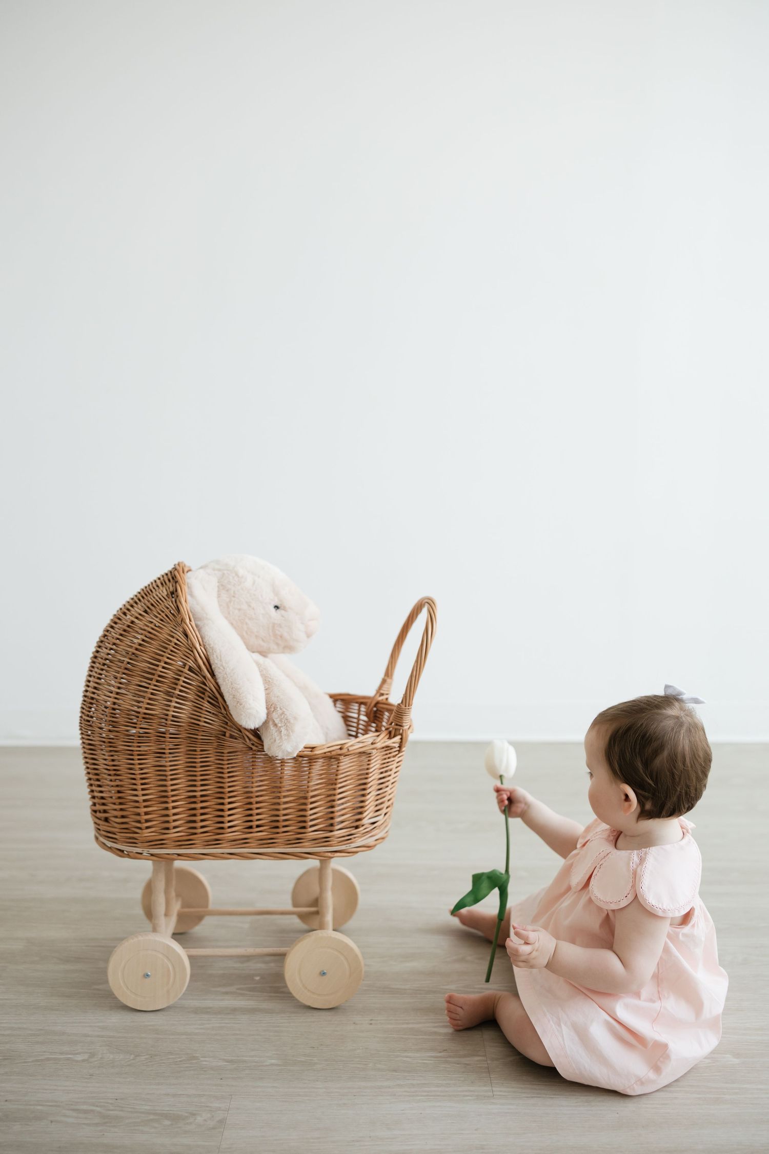 Baby girl in pink dress sits on floor holding white flower beside wicker doll pram with stuffed bunny.