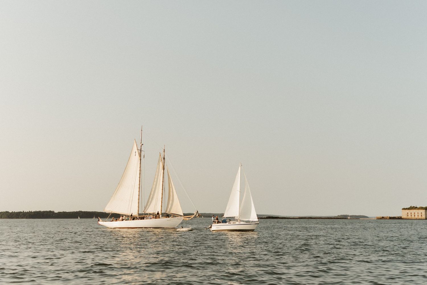 Two white sailboats gliding on calm blue water under a clear sky near a historic waterfront.