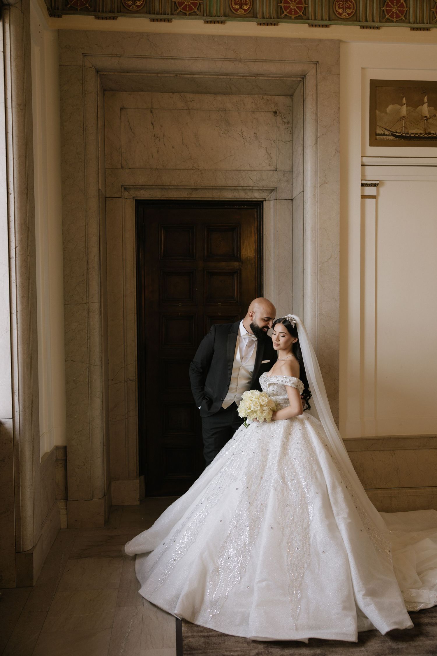 A bride in a luxurious white lace ballgown poses gracefully in an elegant architectural setting.
