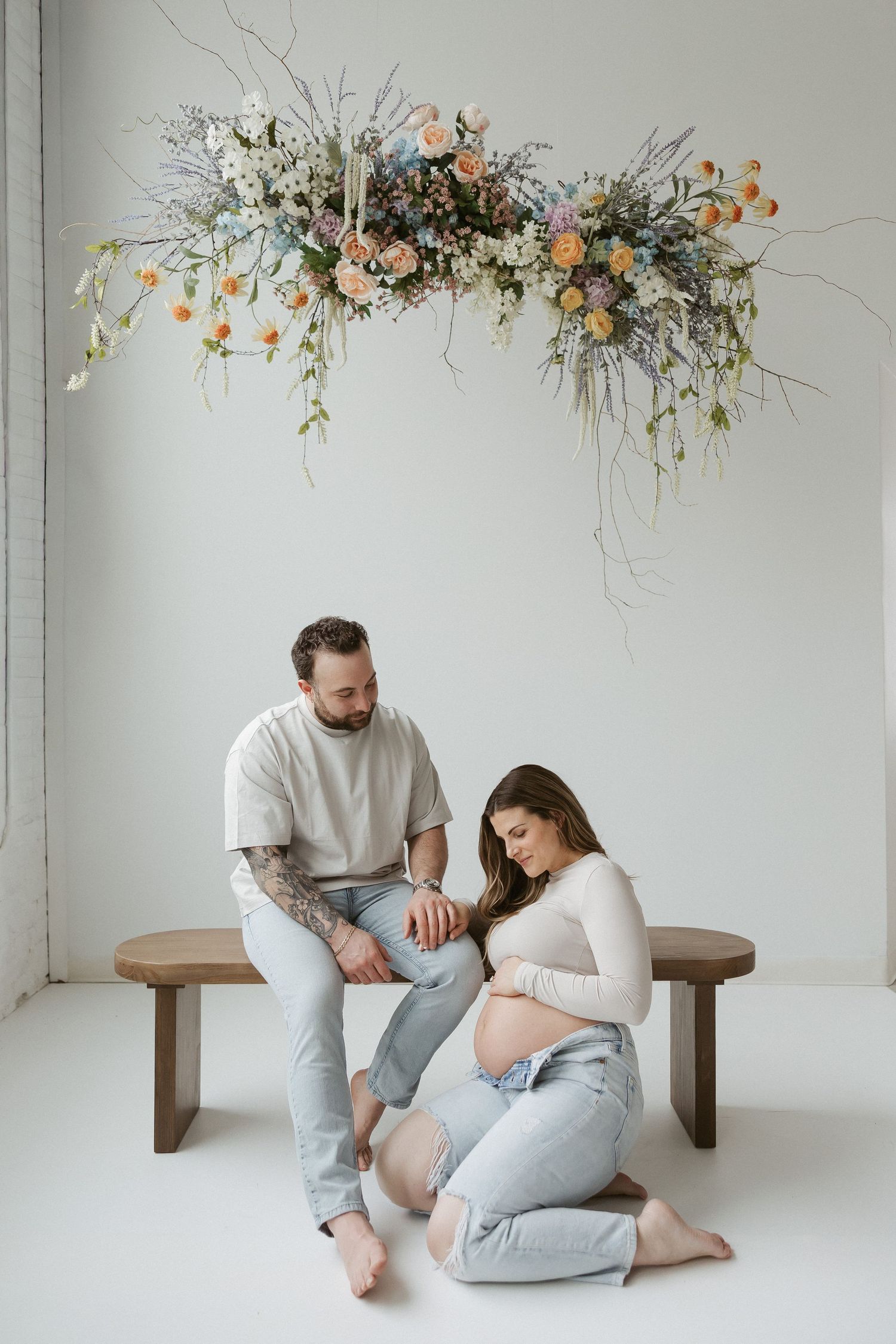 Pregnant couple in a bright studio, man seated on bench beside woman kneeling, floral arrangement overhead.