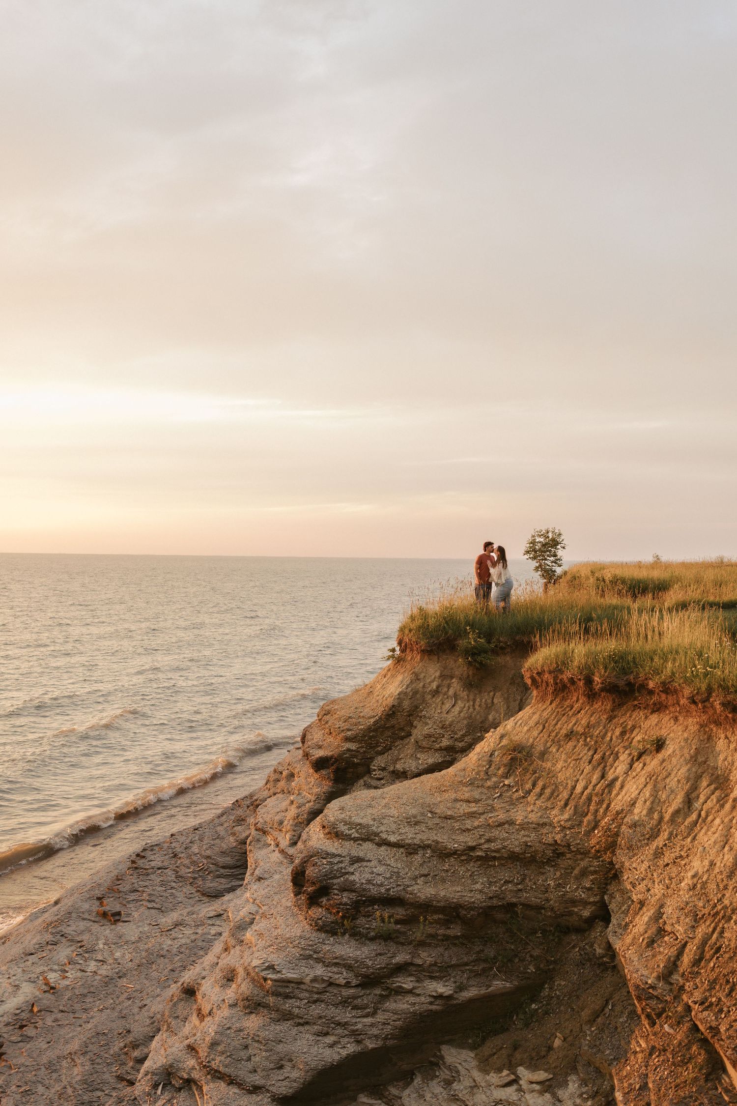 Couple embracing on a grassy cliff edge at sunset overlooking a calm sea with a warm glowing sky.