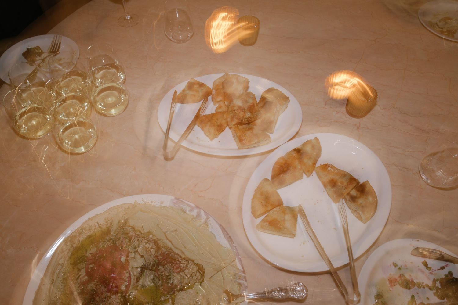 Elegant dinner table setting with appetizer plates, tongs, wine glasses, and candles in warm ambient lighting.