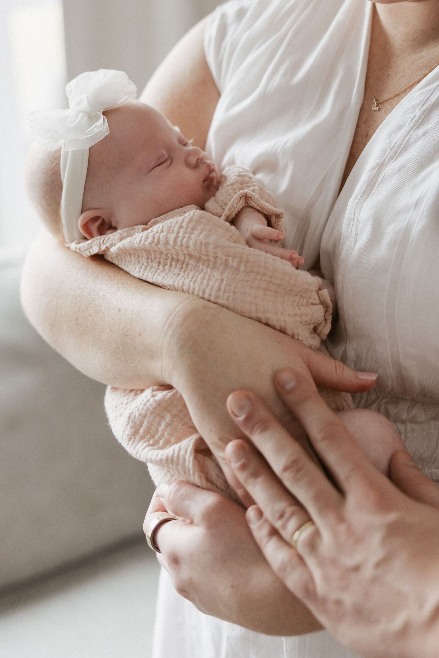 A mother in white gently cradles her sleeping newborn baby wrapped in a soft pink muslin cloth with a white bow headband.