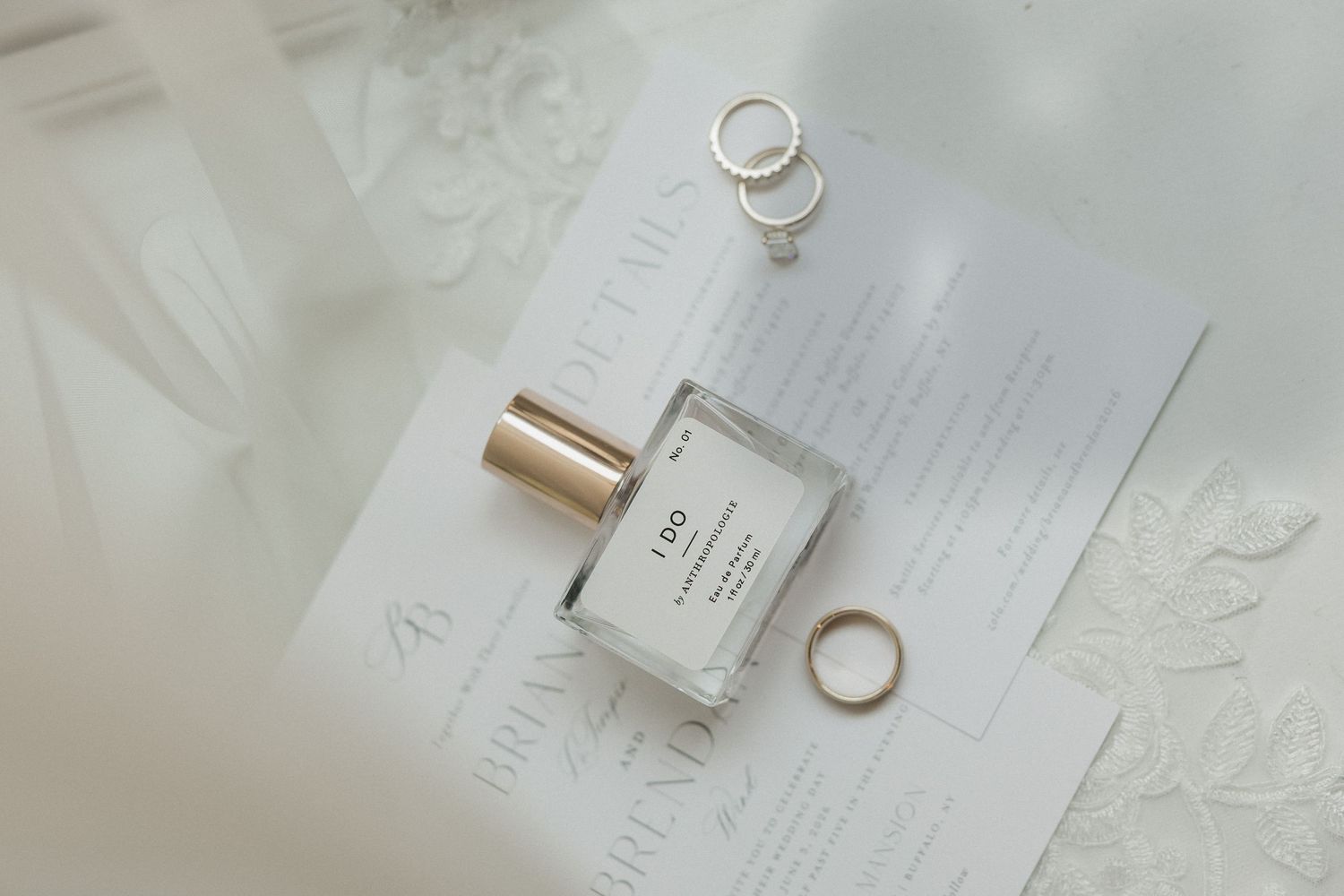 Elegant wedding flat lay with perfume bottle, gold rings, and floral stationery on white fabric.
