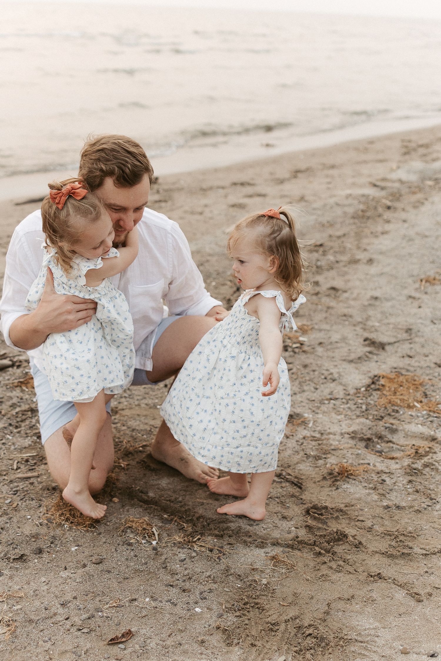 Two people in white outfits sit together on a sandy beach examining seashells and treasures from the shore.
