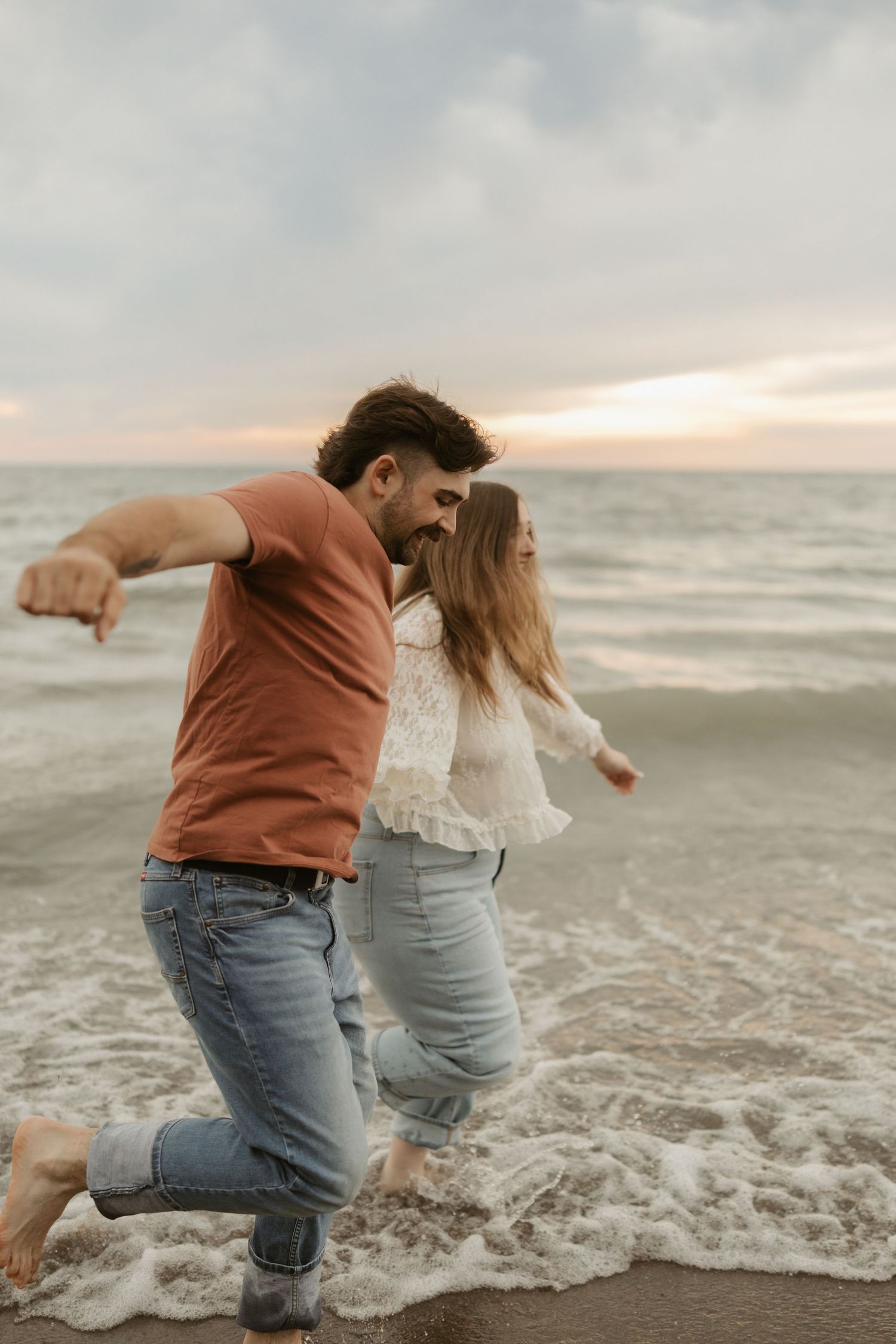 Couple playing in ocean waves at sunset, wearing jeans and casual tops, laughing and splashing on sandy beach.