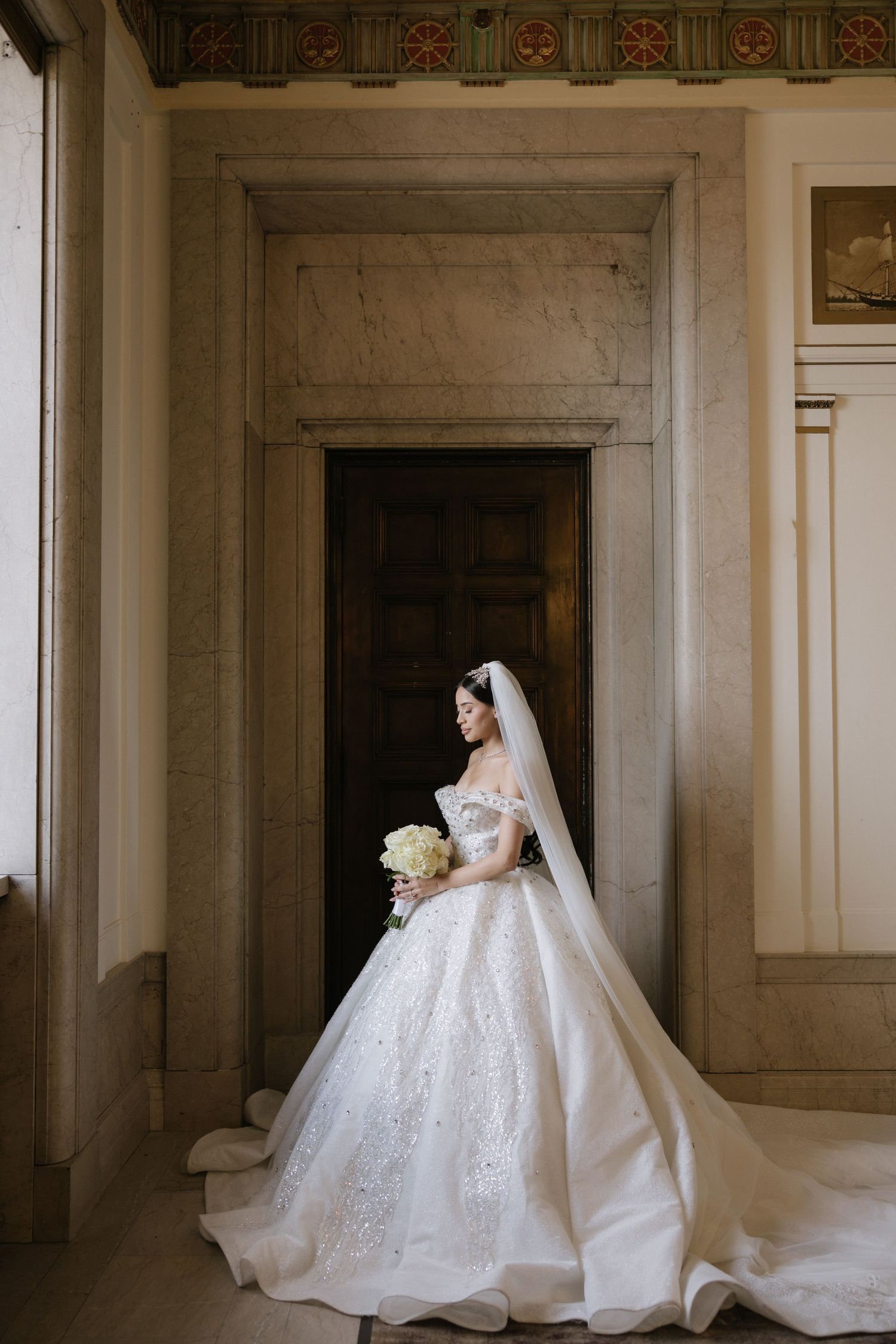 A bride in an elegant white ball gown with long train poses gracefully in a grand marble hallway.