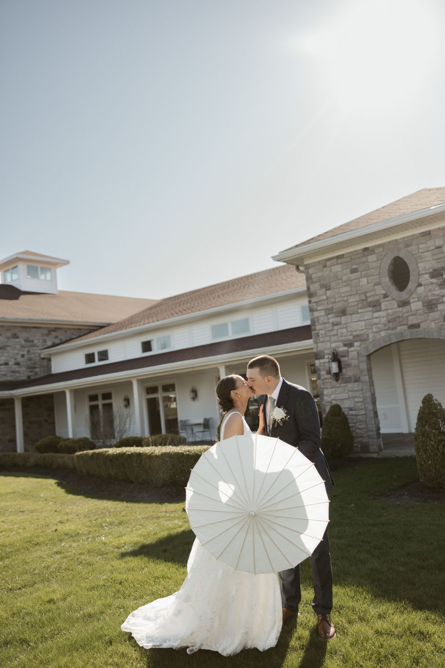 Bride and groom kissing outdoors holding a white parasol in front of an elegant stone wedding venue.