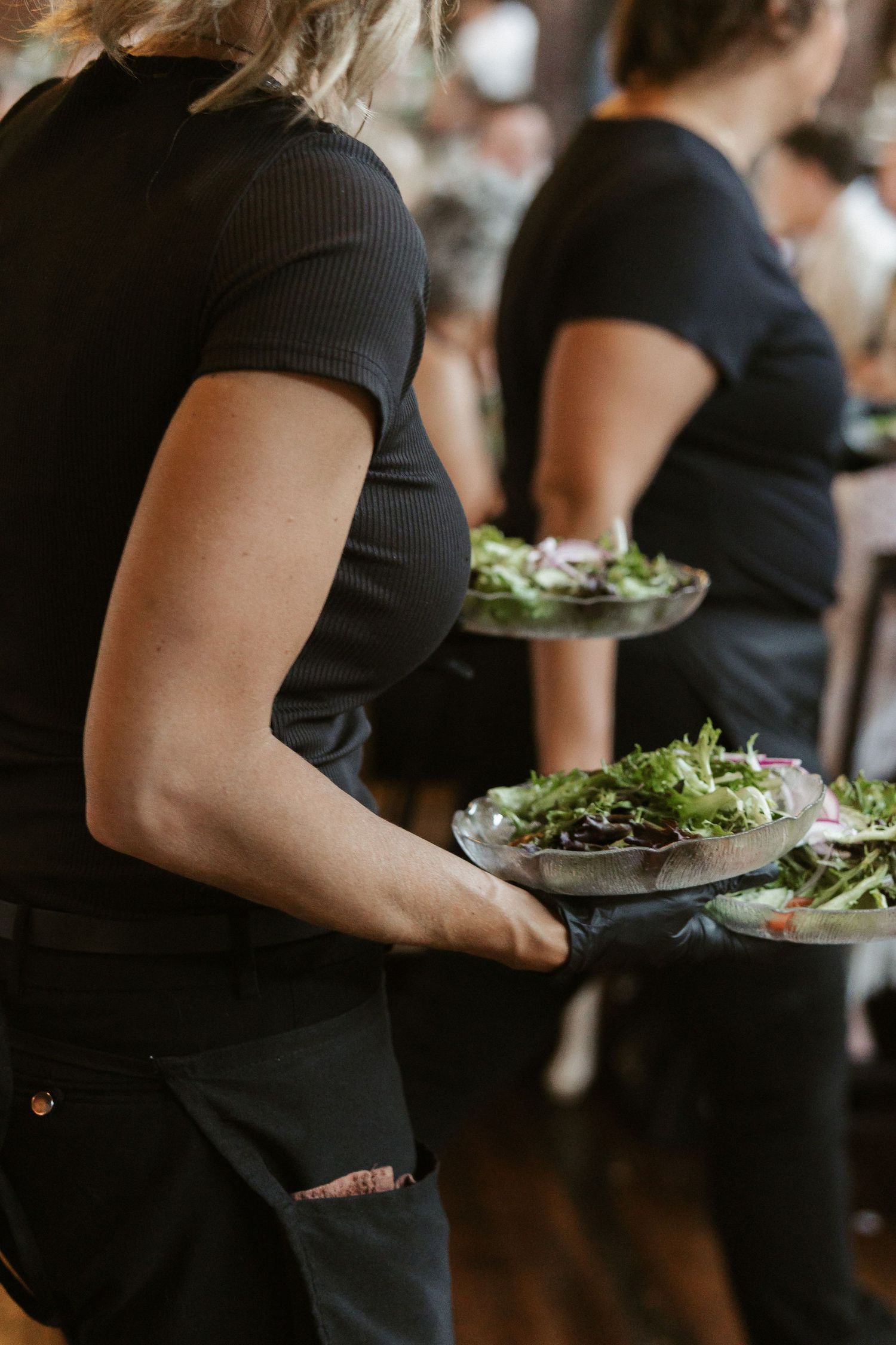 Line of servers in black attire carrying plates of fresh garden salads at an elegant event.