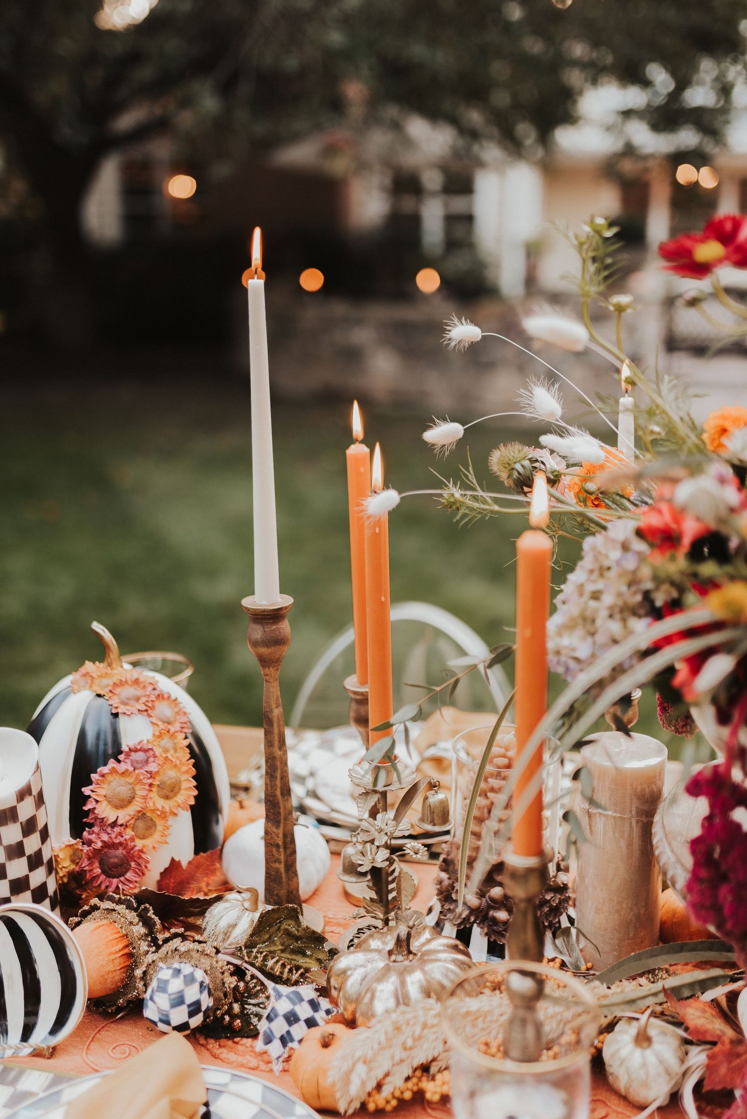 Elegant fall table setting with lit candles, pumpkins, and floral decorations at dusk with bokeh lights in background.