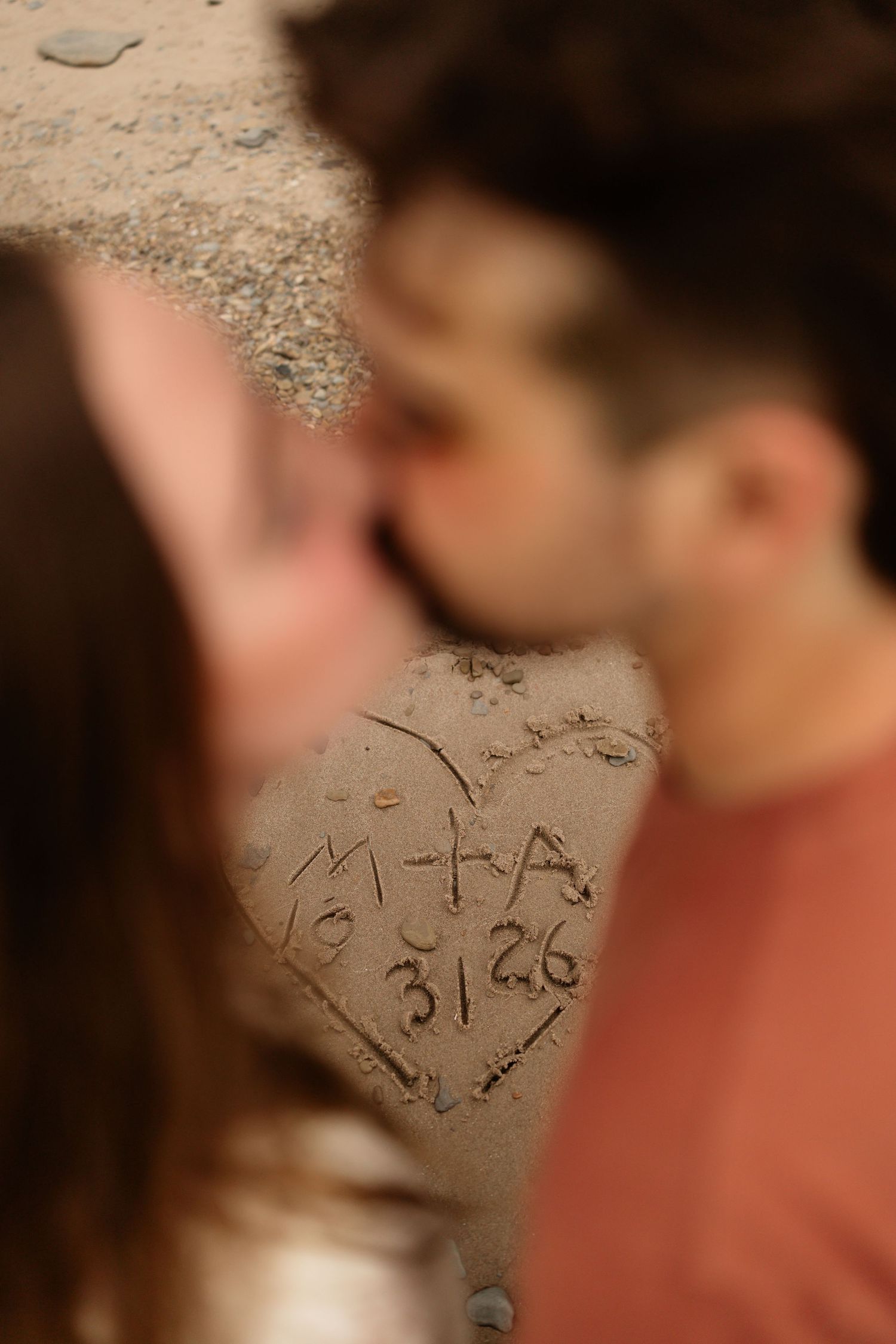Couple kissing behind a heart drawn in sand with initials M+A and date 3/26 at a beach.