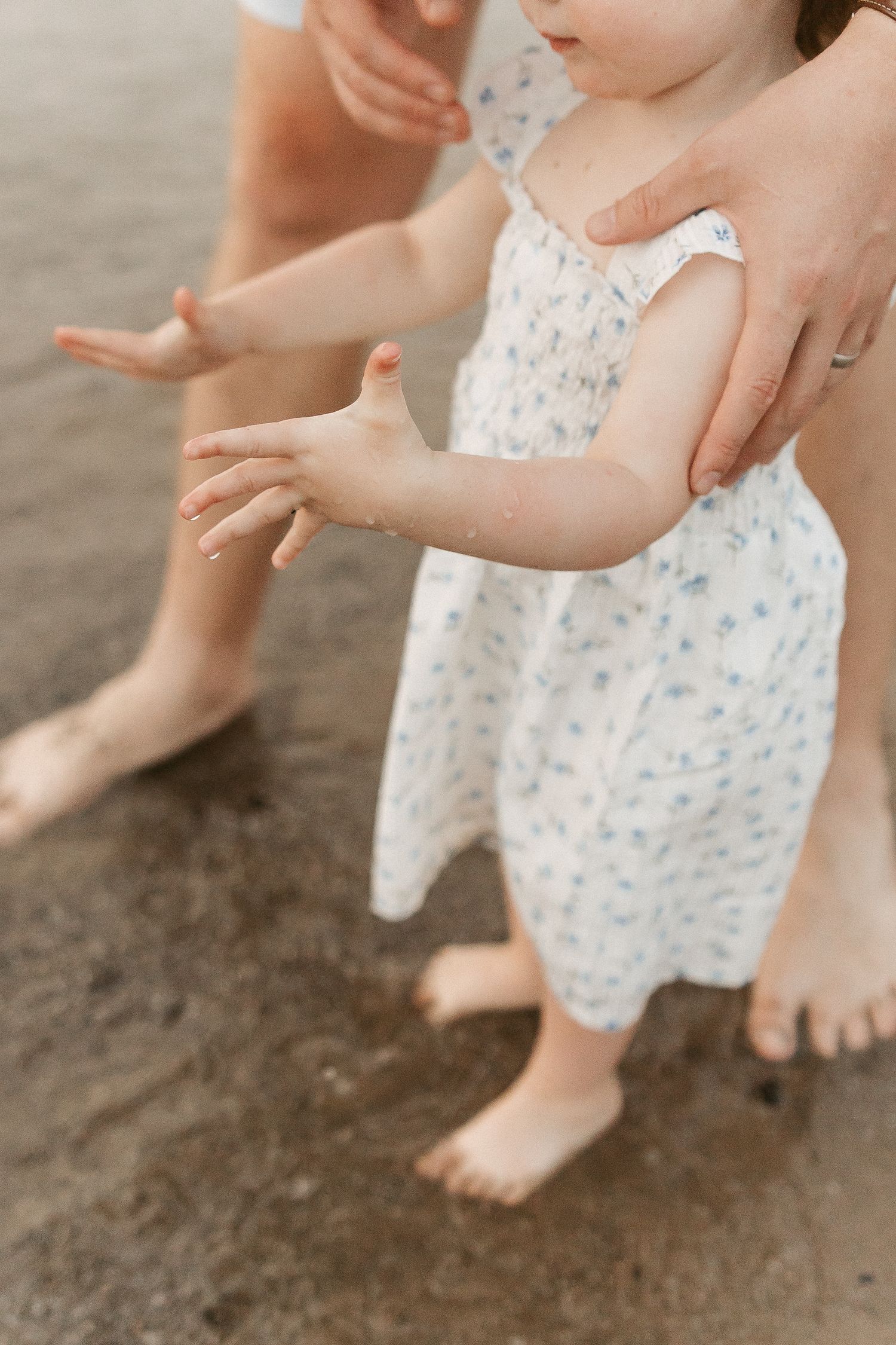 A toddler in a white floral dress stands barefoot on wet sand, supported by an adult's hands at the beach.