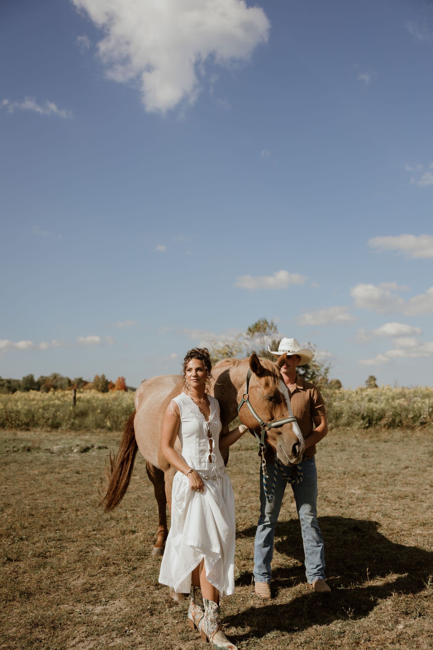 A couple in western attire poses with their horse in a sunny field during a rustic outdoor photoshoot.