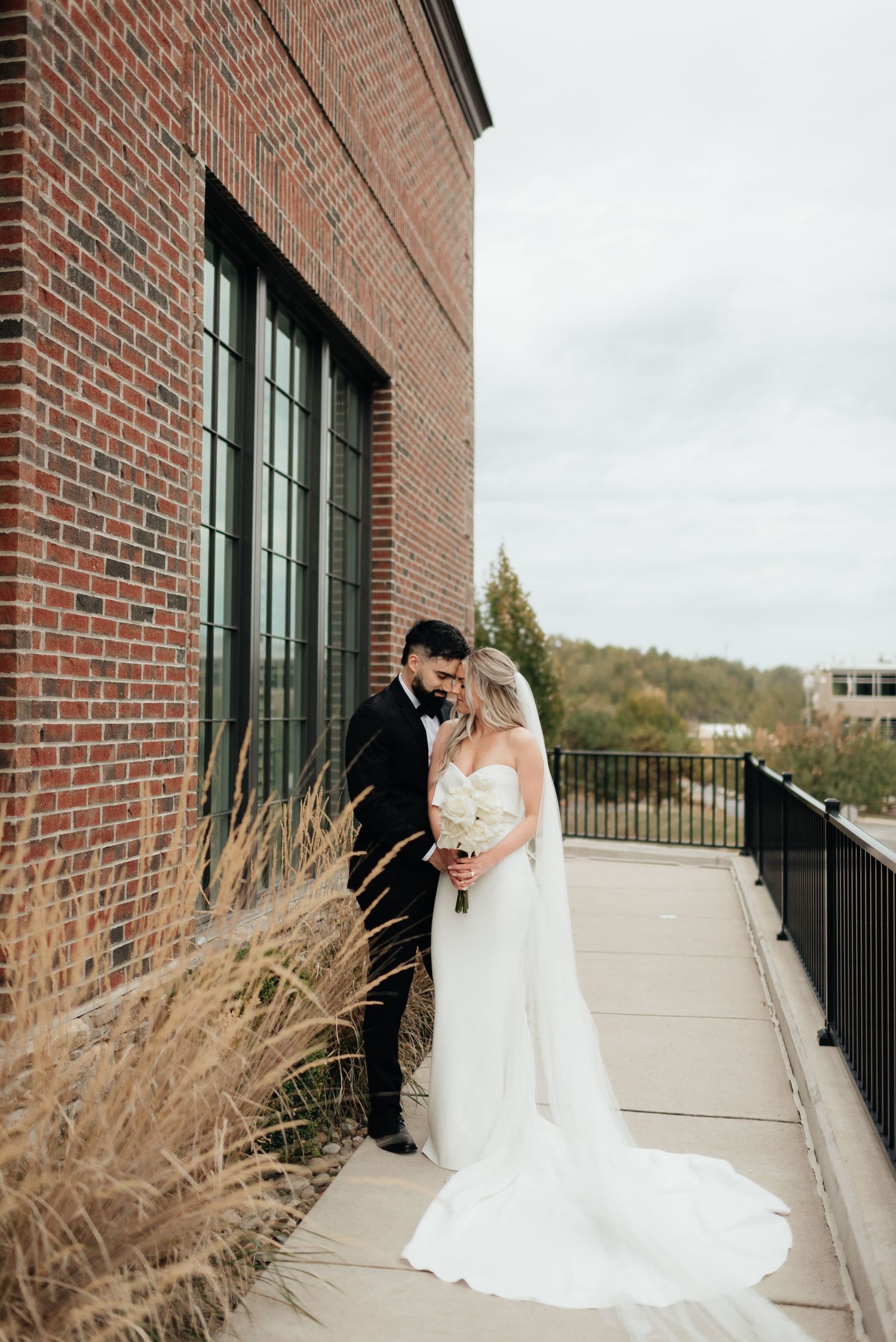 A couple in wedding attire embraces on a brick building balcony with ornamental grasses in autumn.
