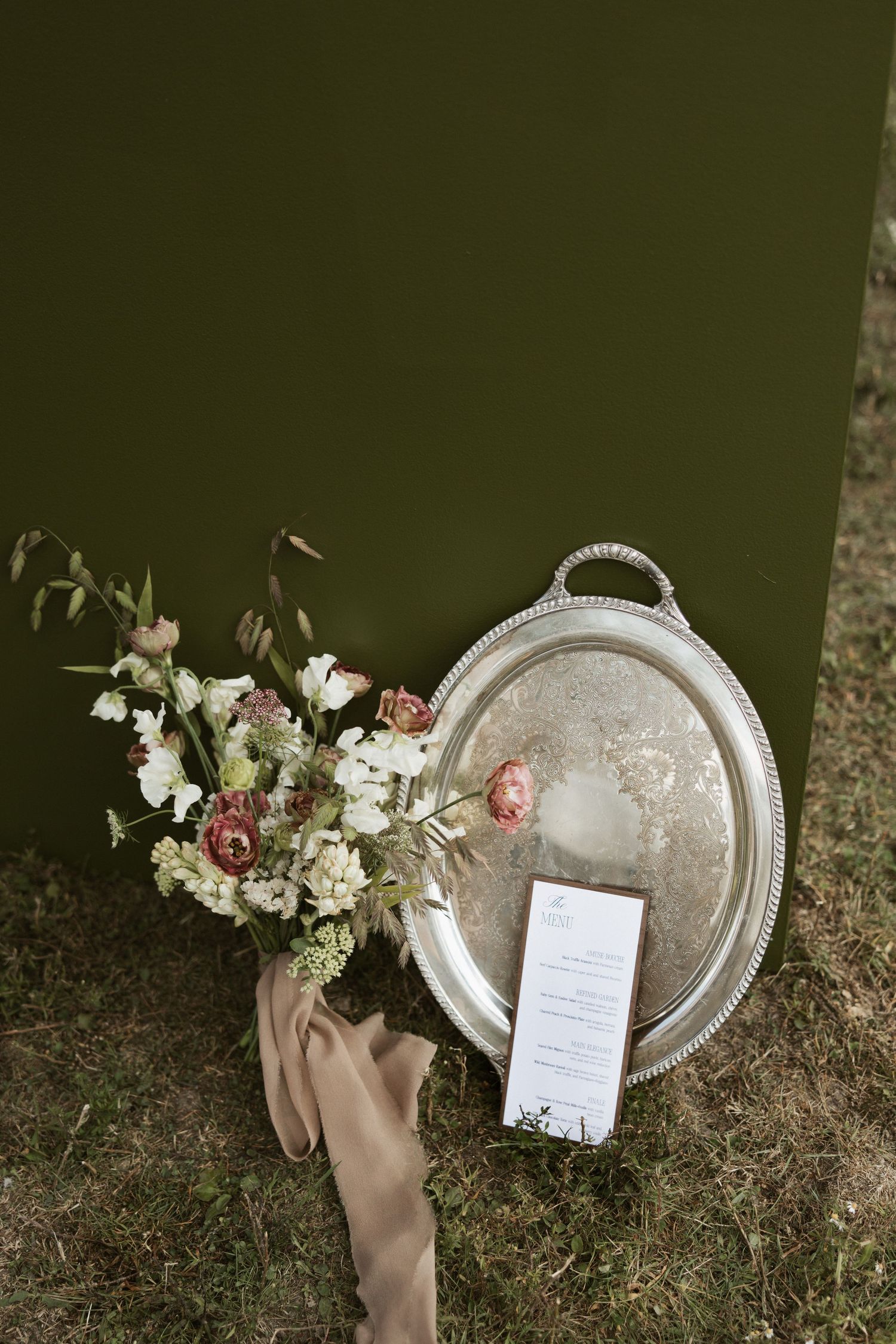A decorative silver platter with a menu card sits on grass alongside elegant floral arrangements in neutral tones.