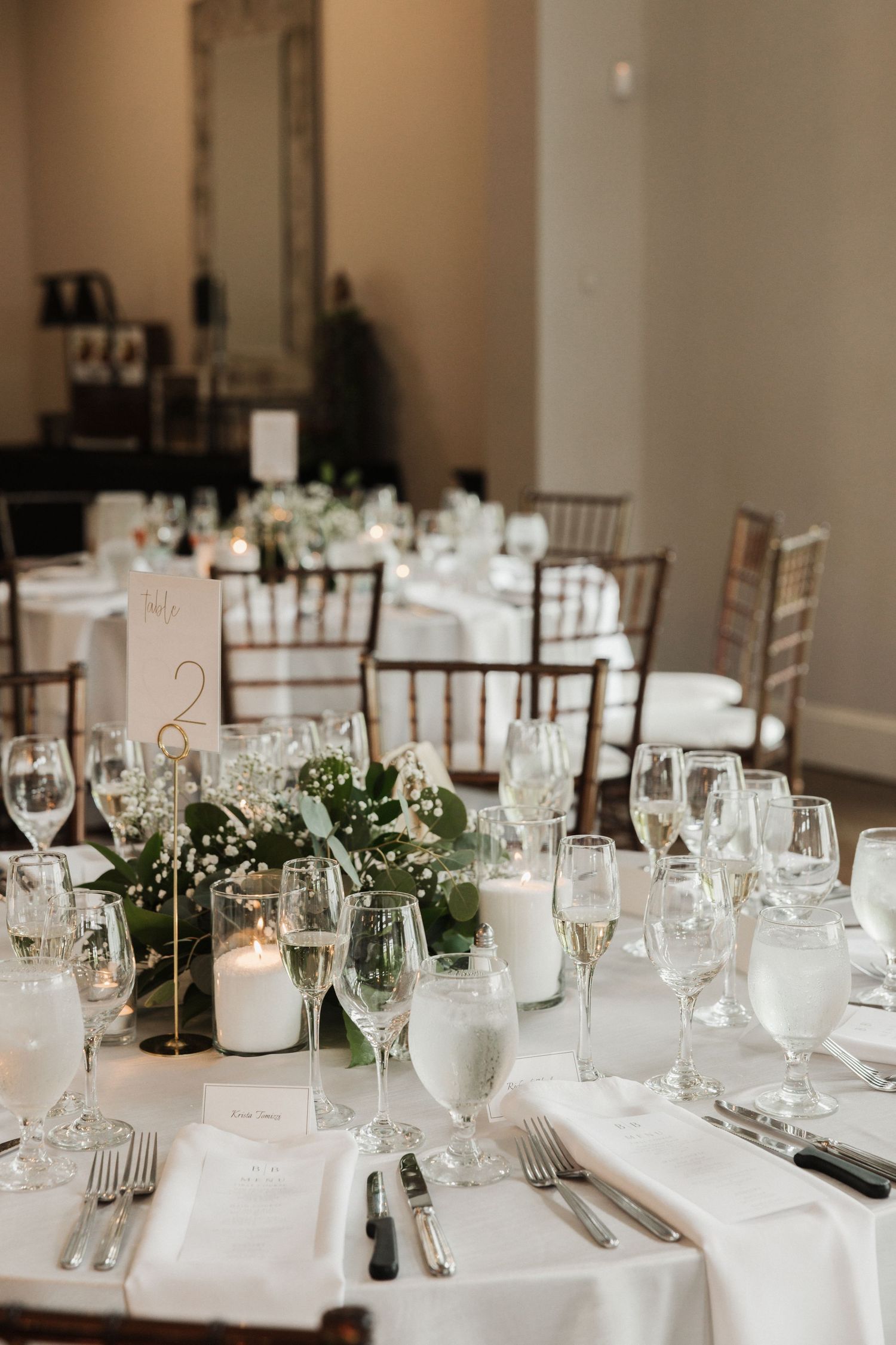 Elegant wedding reception table with white linens, greenery centerpieces, candles, crystal glassware, and gold table numbers.