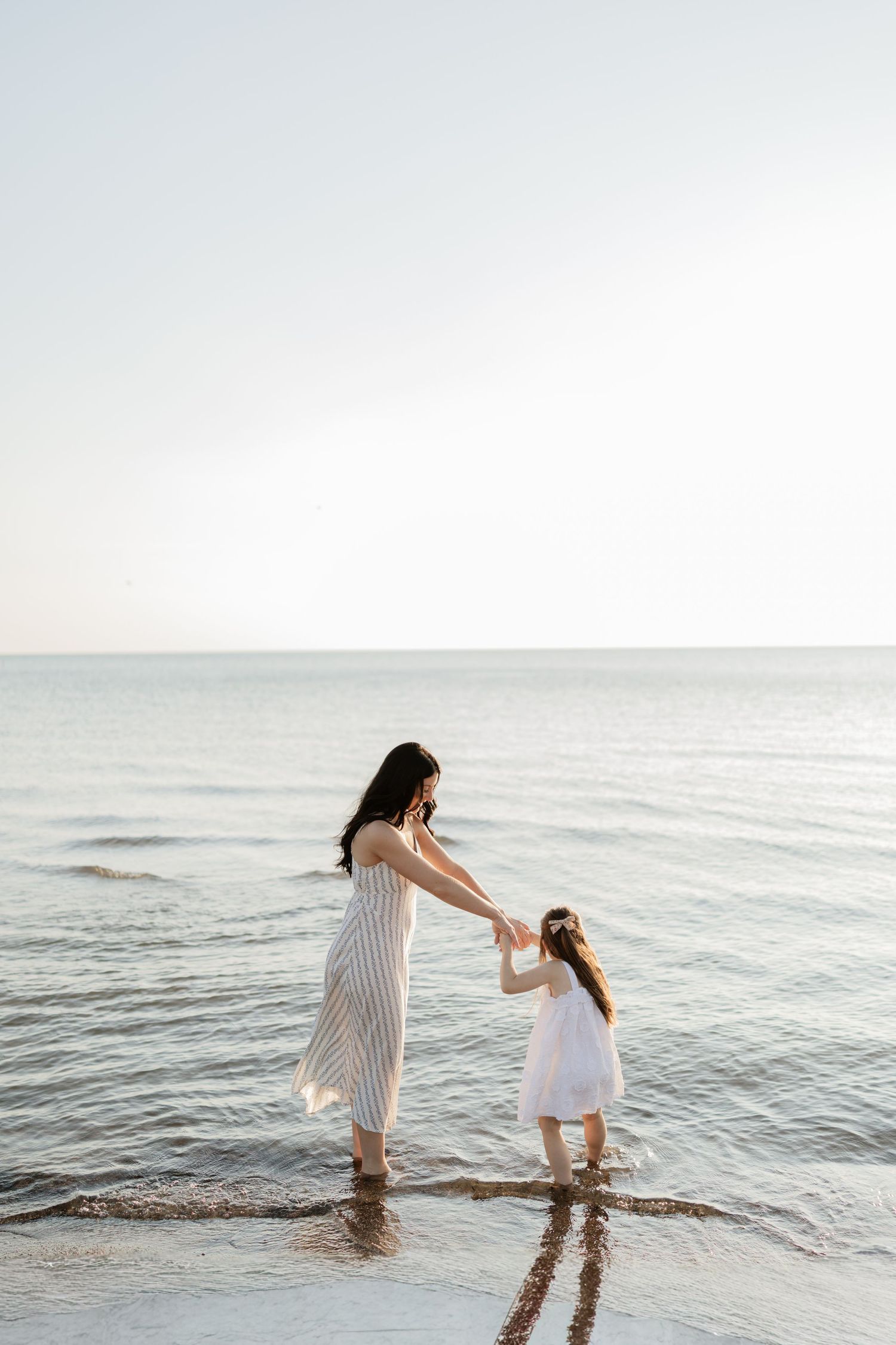 A mother and daughter in white dresses hold hands while wading in calm ocean waves at the beach.