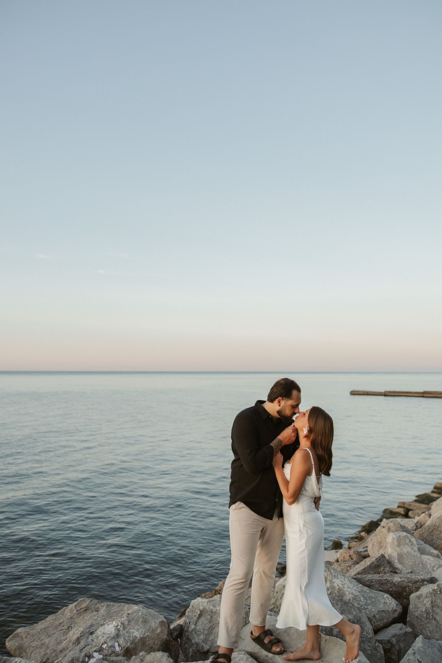 A couple shares a romantic kiss on rocky coastal breakwater during sunset by the ocean.