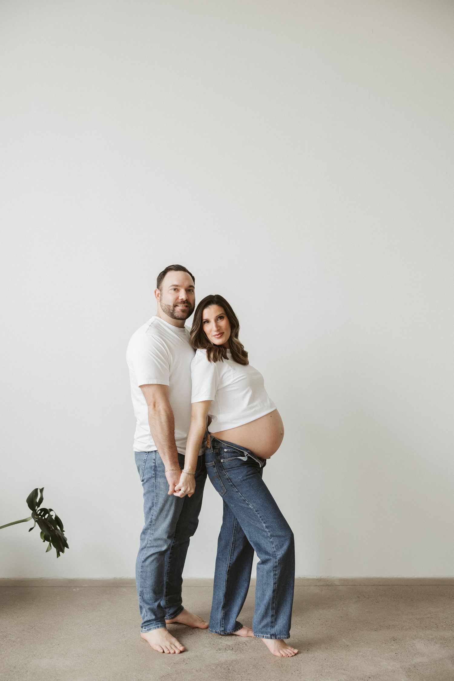 Couple in white tops and jeans posing for maternity photo, woman showing baby bump, holding hands against white wall.