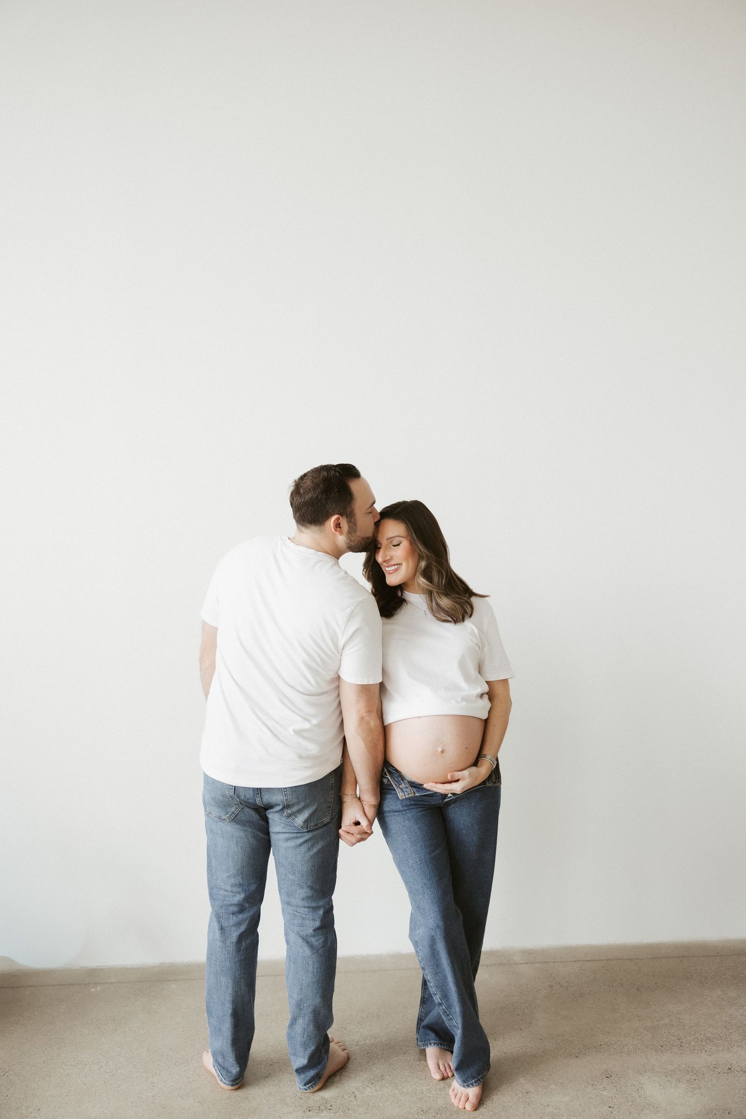 Couple in white tees and jeans sharing a tender moment, man kissing pregnant woman's cheek in a bright studio.