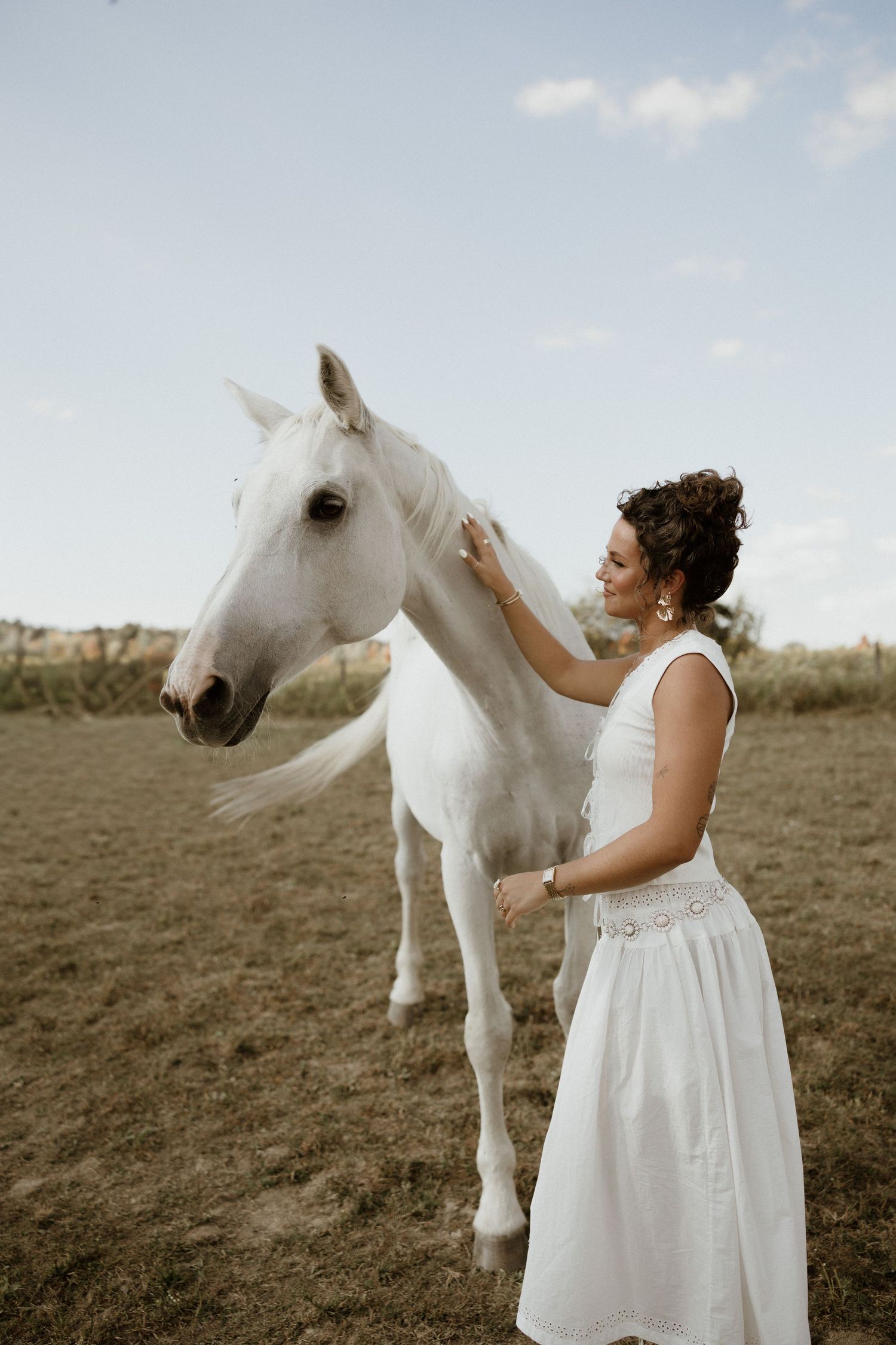 A person in a white dress stands in a field with a majestic white horse during a dreamy outdoor photoshoot.