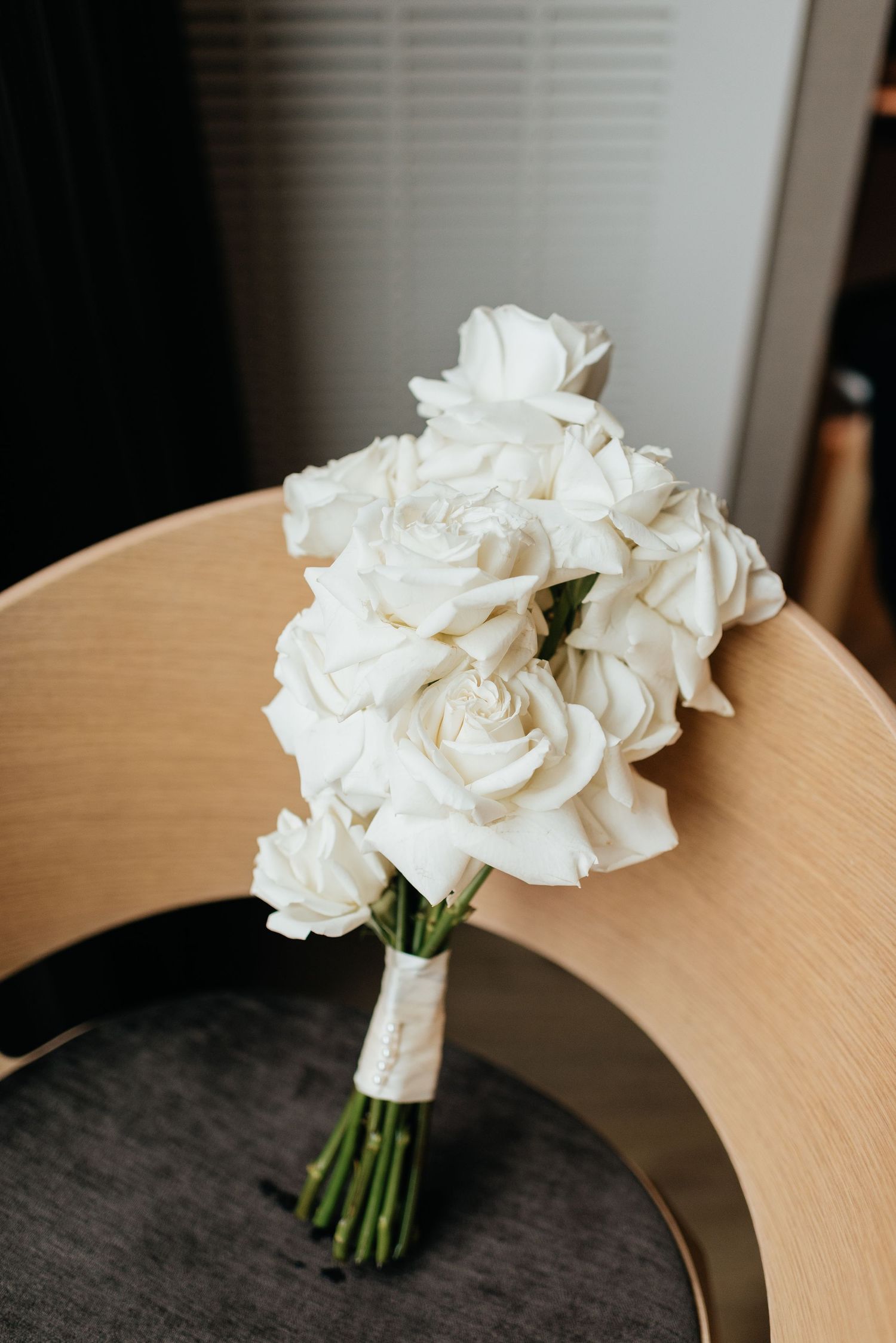 A classic white rose bridal bouquet wrapped in ribbon sits on a wooden chair against a dark background.