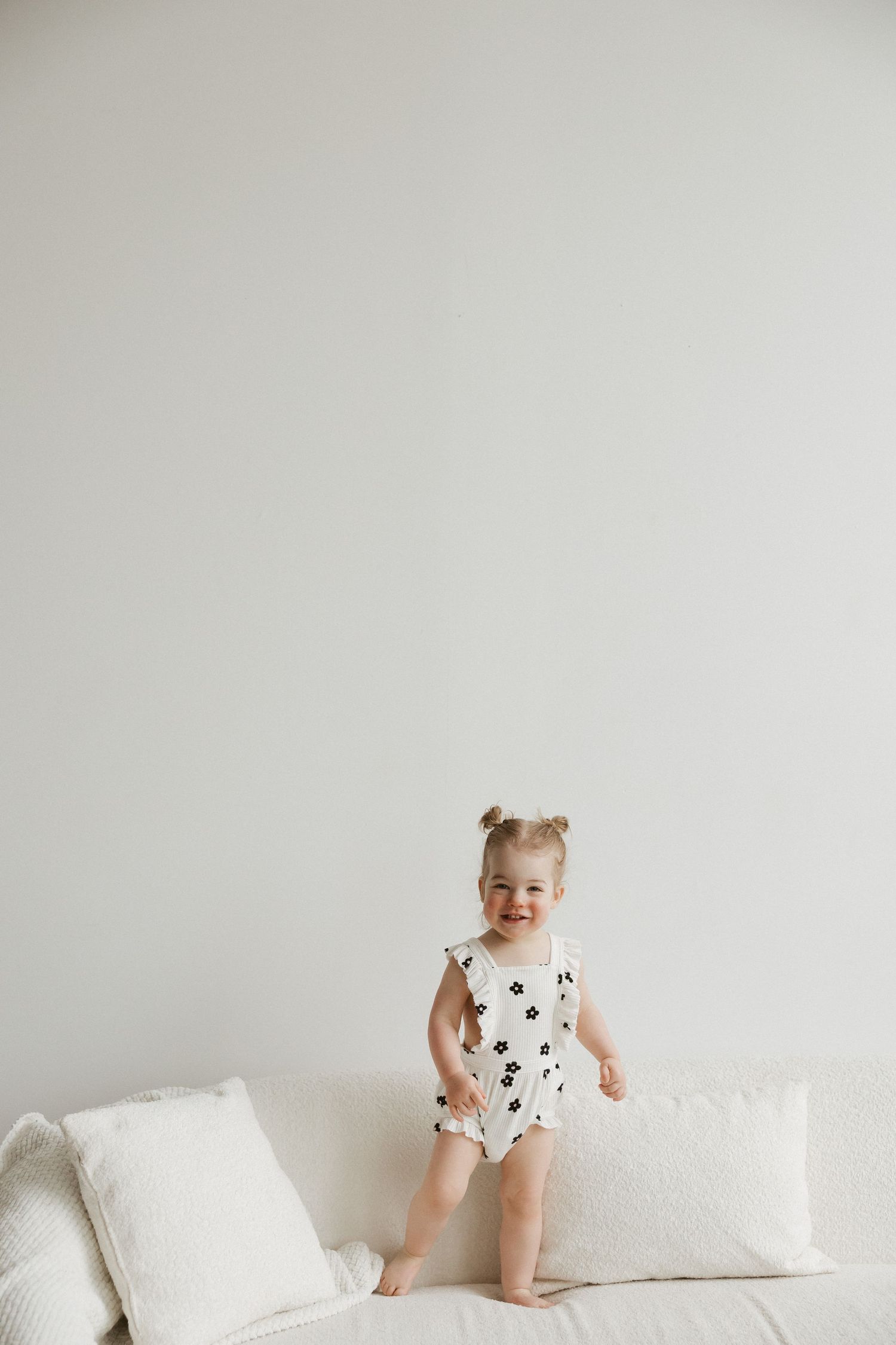 Smiling toddler girl in white floral romper standing on a white sofa against a light gray wall.