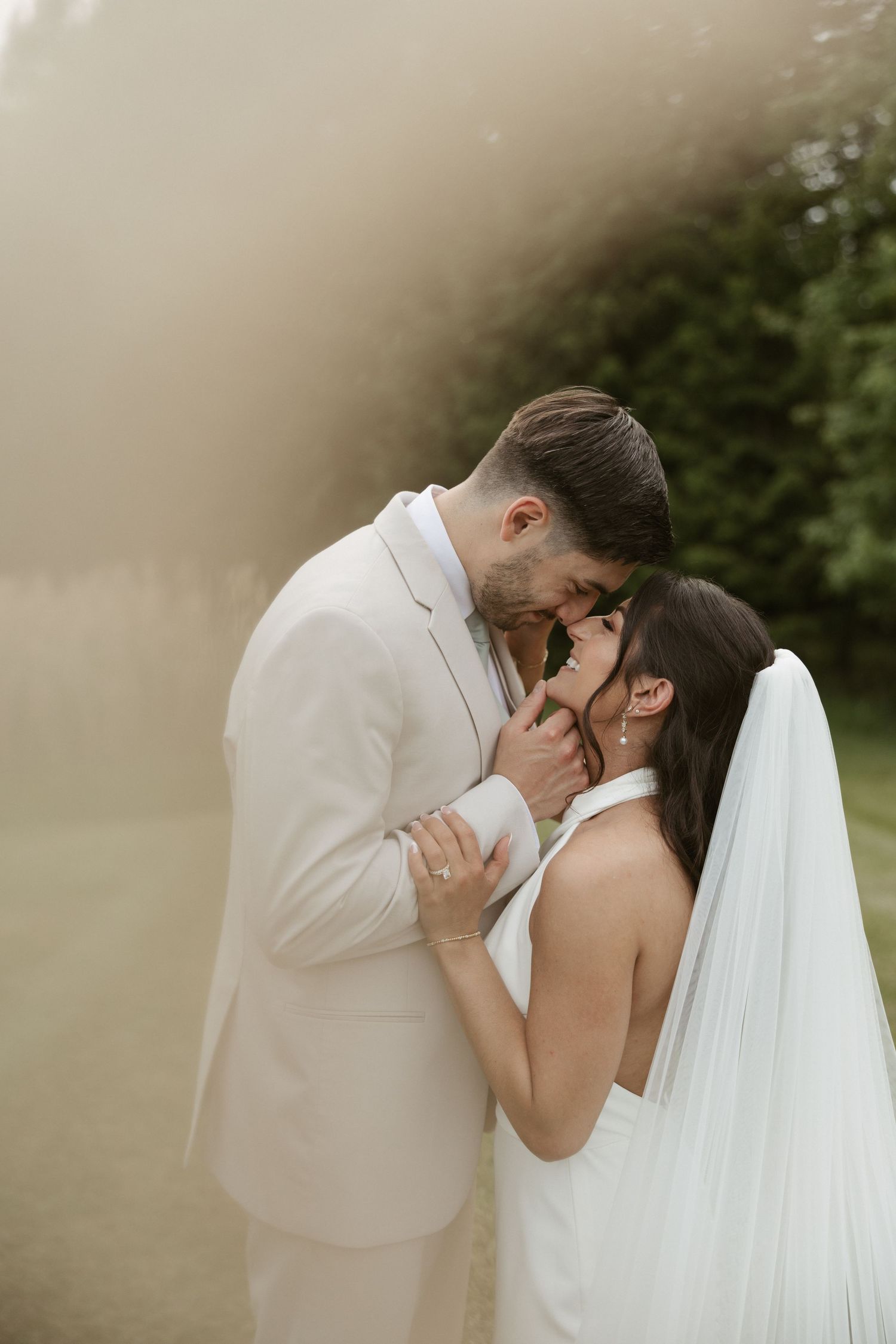 A bride and groom share an intimate moment, nearly kissing outdoors in soft golden light, dressed in white wedding attire.
