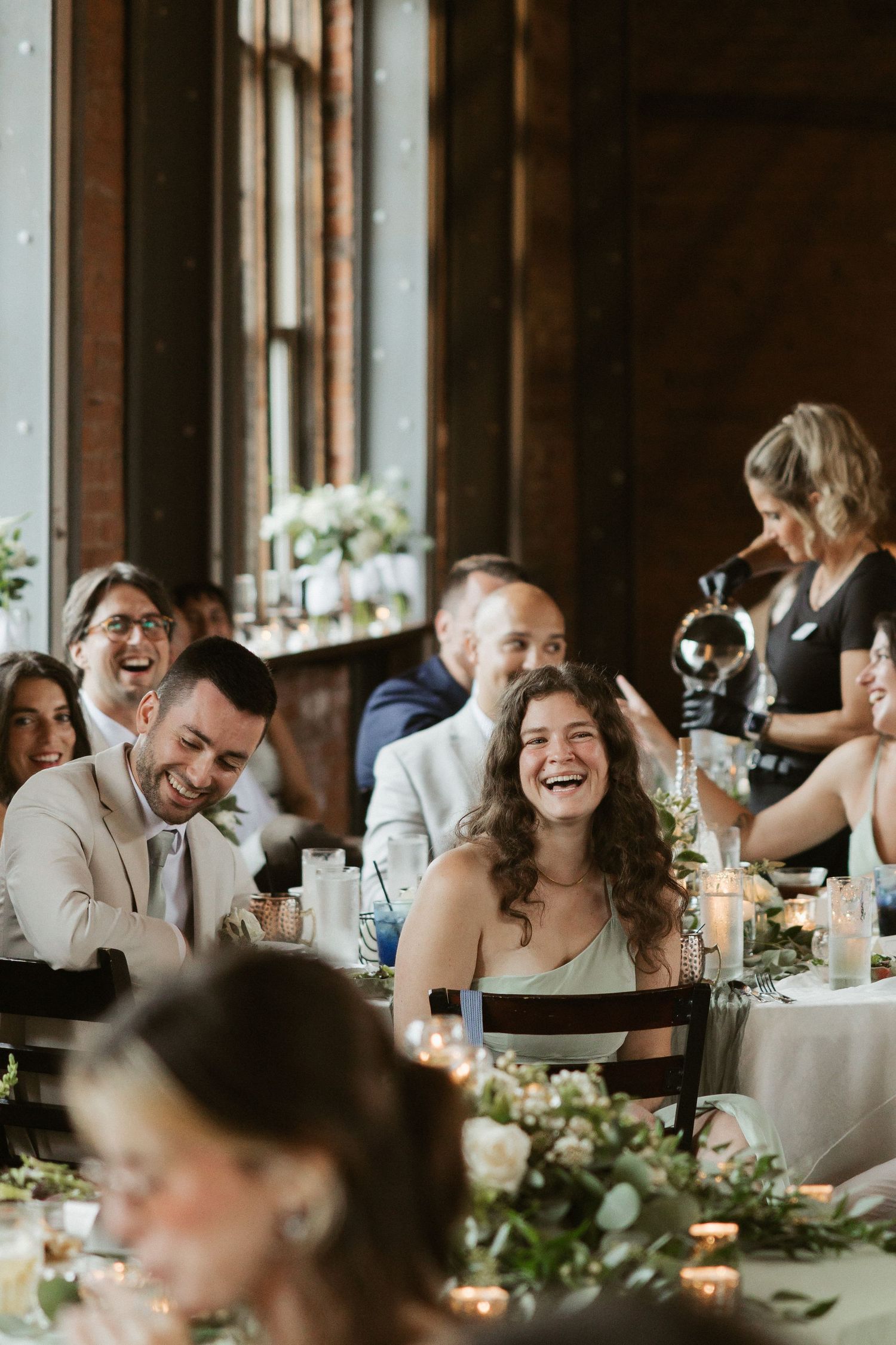 Wedding reception guests share laughter and joy at long banquet table decorated with greenery garlands and candlelight.