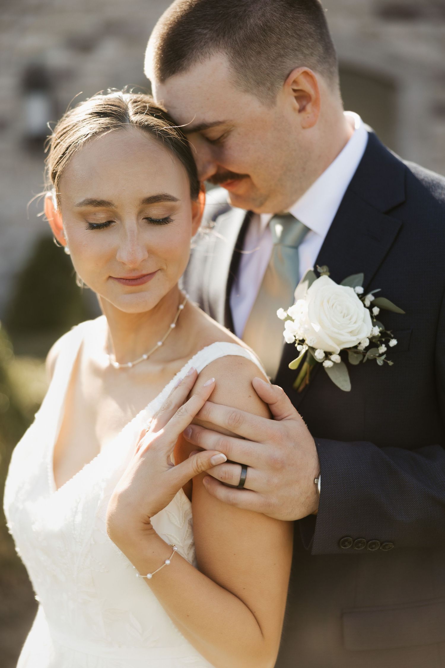 Bride and groom share tender moment, she wears white dress and ring, he wears navy suit with white rose boutonniere.