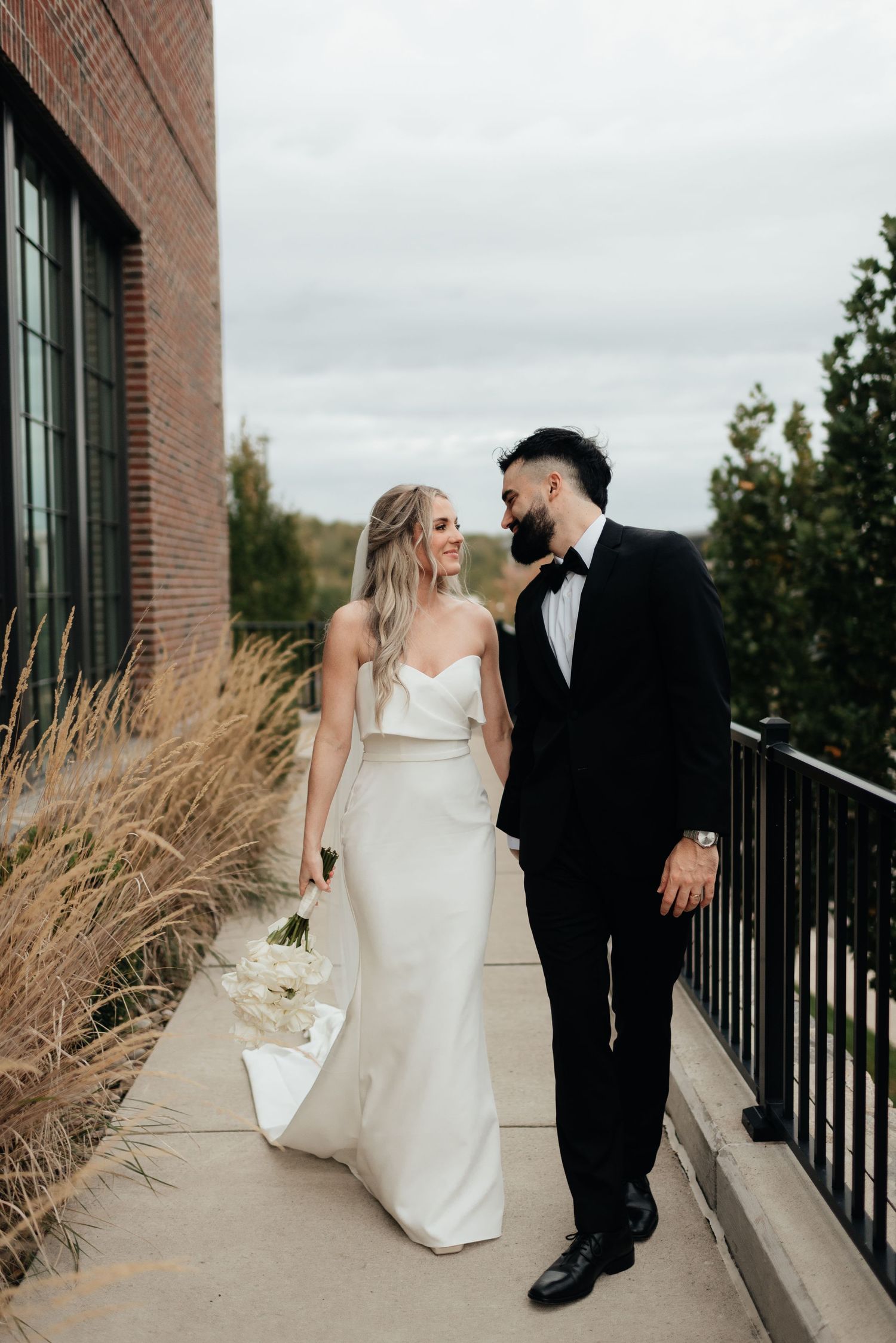 A couple in wedding attire walk together on a concrete path next to ornamental grasses and a brick building.