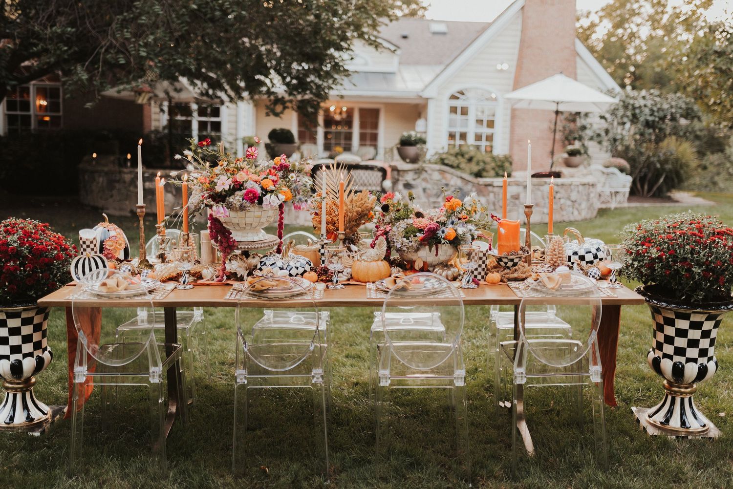 Elegant outdoor dinner table setting with ghost chairs and fall-themed decor in a backyard garden with house in background.