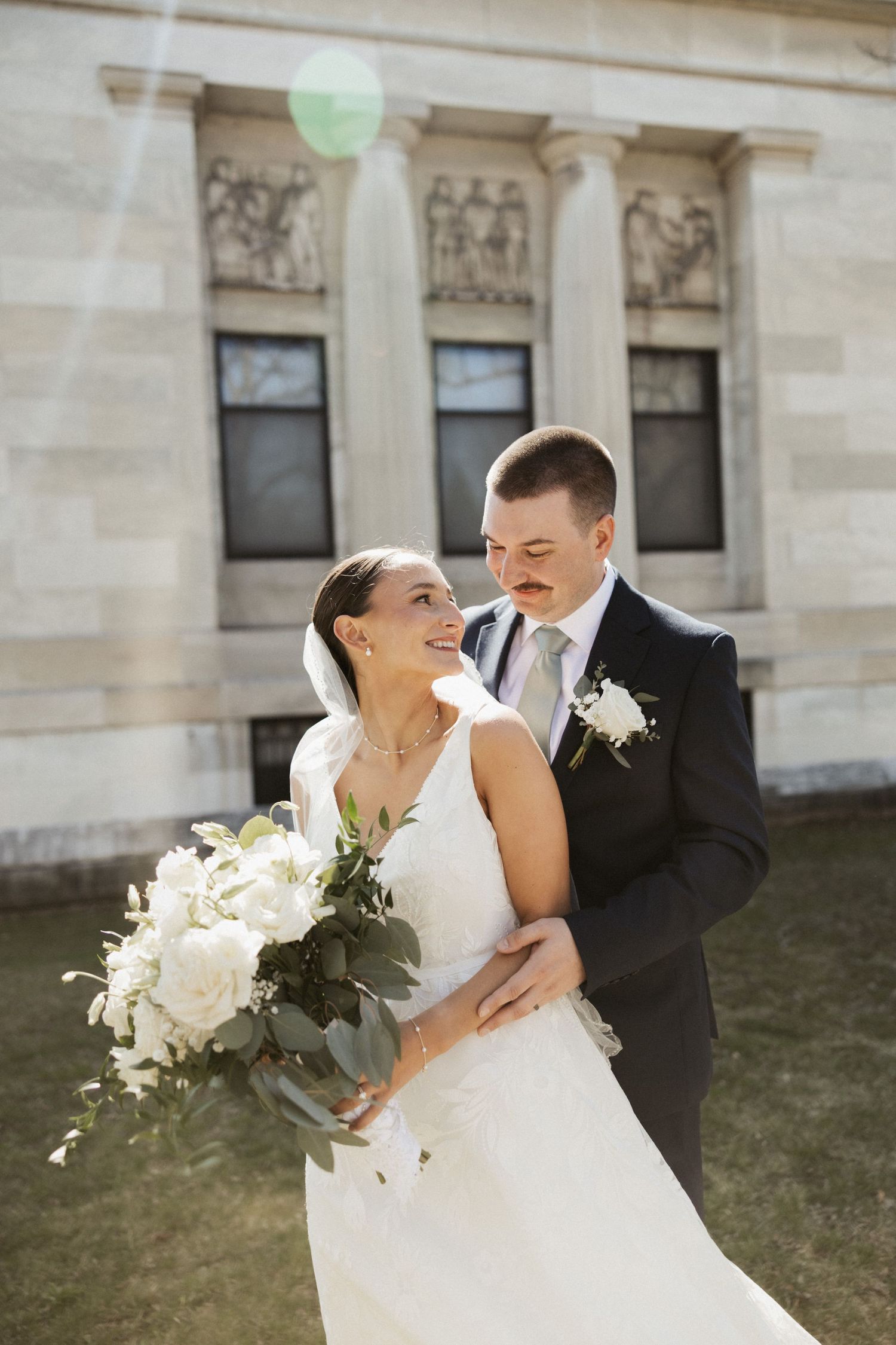 Bride and groom embrace outdoors in front of a classical stone building, bride holding white floral bouquet in wedding attire.