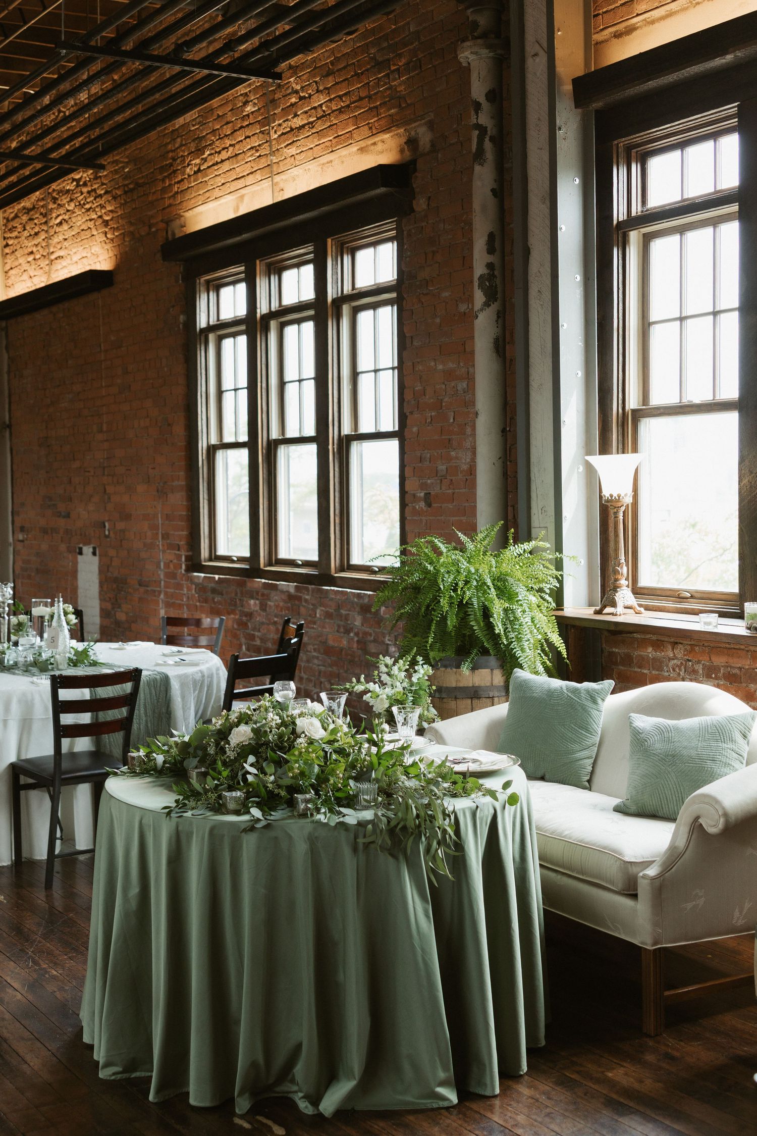 An elegant reception venue with exposed brick walls, tall windows, and sage green tablecloths adorned with greenery.