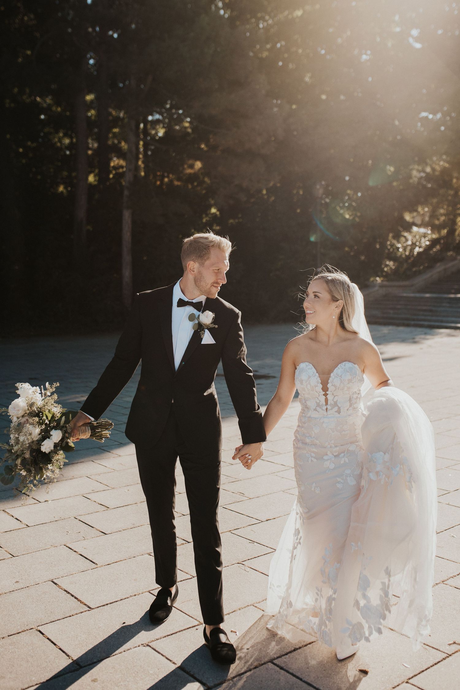 A couple walking together on a stone path in sunlight, the bride in a lace gown and groom in classic black tuxedo.