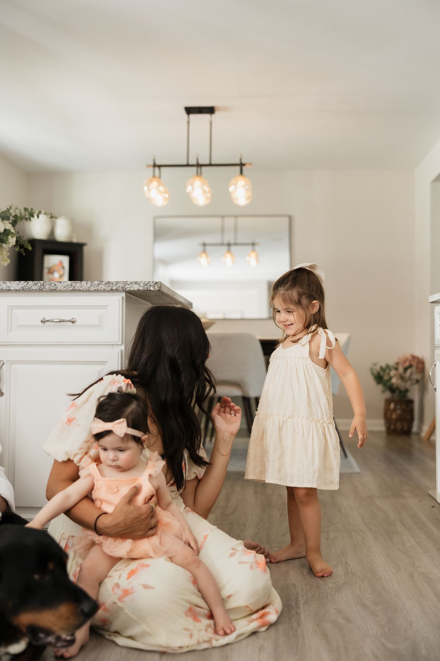 A mother sits on the floor with her baby and toddler daughter in a bright modern kitchen, with a dog nearby.