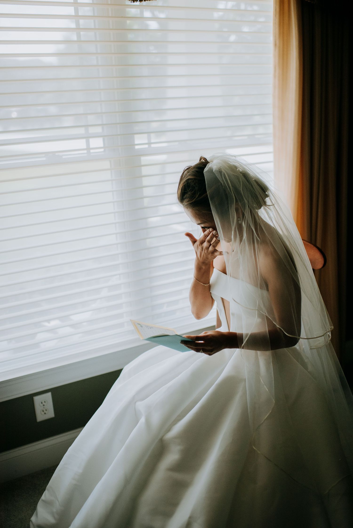 A bride in a white wedding gown sits by a window with venetian blinds while reading from a book.