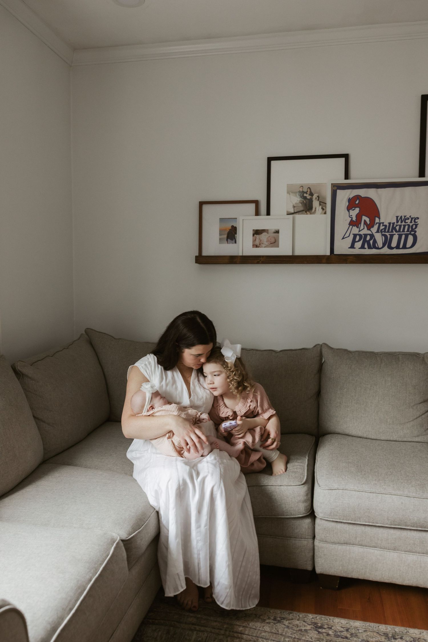 A mother in a white dress sits on a gray sectional sofa with her two young children in a cozy living room.