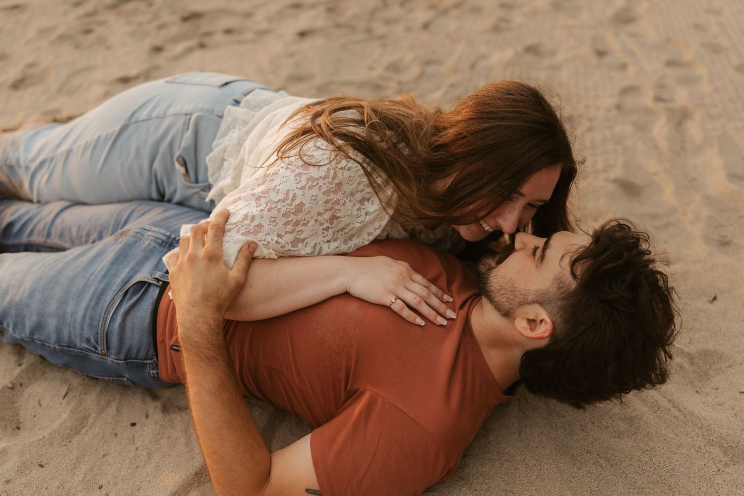 Couple sharing a romantic kiss while lying on sandy beach, woman in lace top leaning over man in rust shirt.