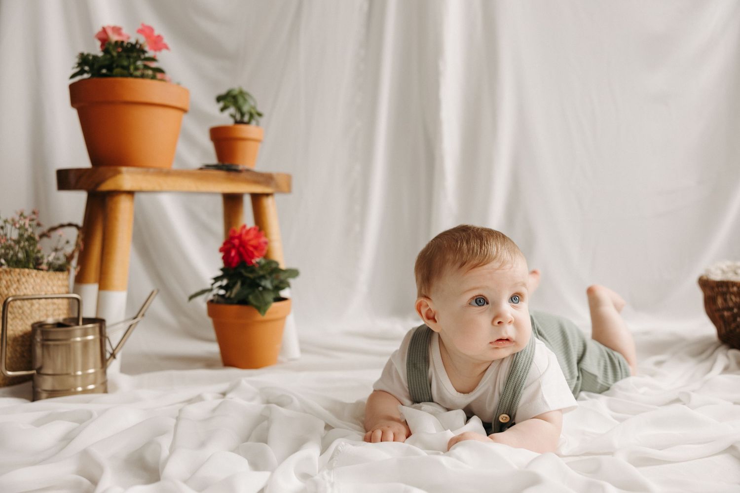 Baby in light green outfit lying on white fabric with terracotta potted plants on wooden shelf in background.
