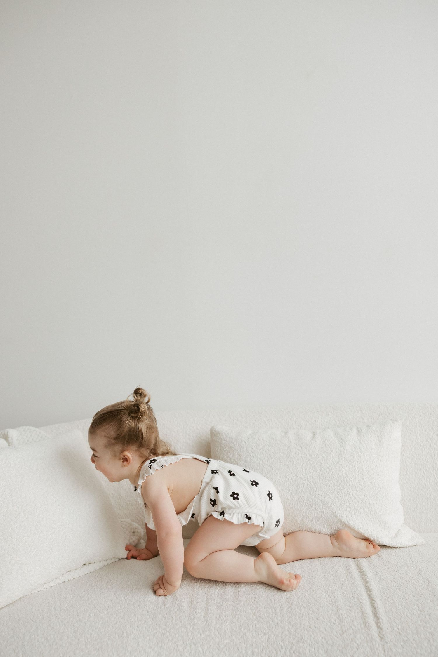 Baby girl in floral romper crawling on white sofa with cushions in bright minimalist room.