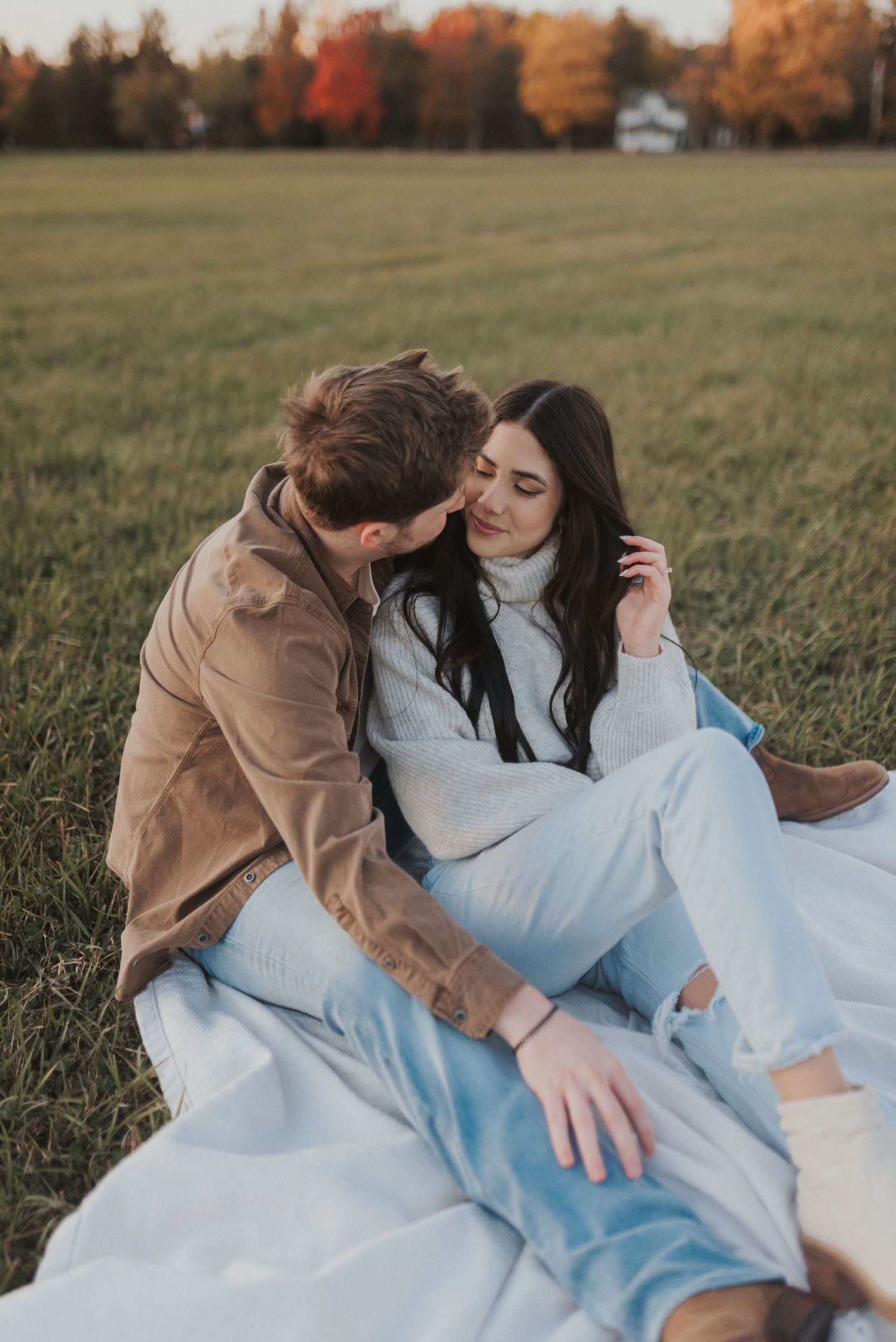 A couple relaxes on a blanket in a grassy field during autumn with colorful fall foliage in the background.