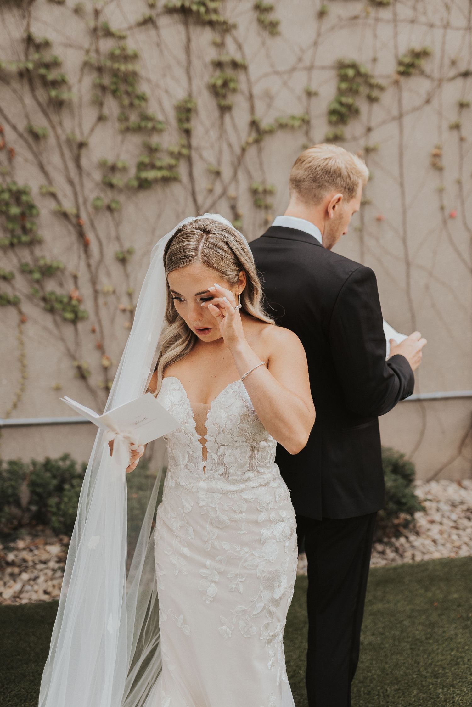 A bride in a lace wedding dress and veil stands back-to-back with a groom in a black suit during their first look moment.