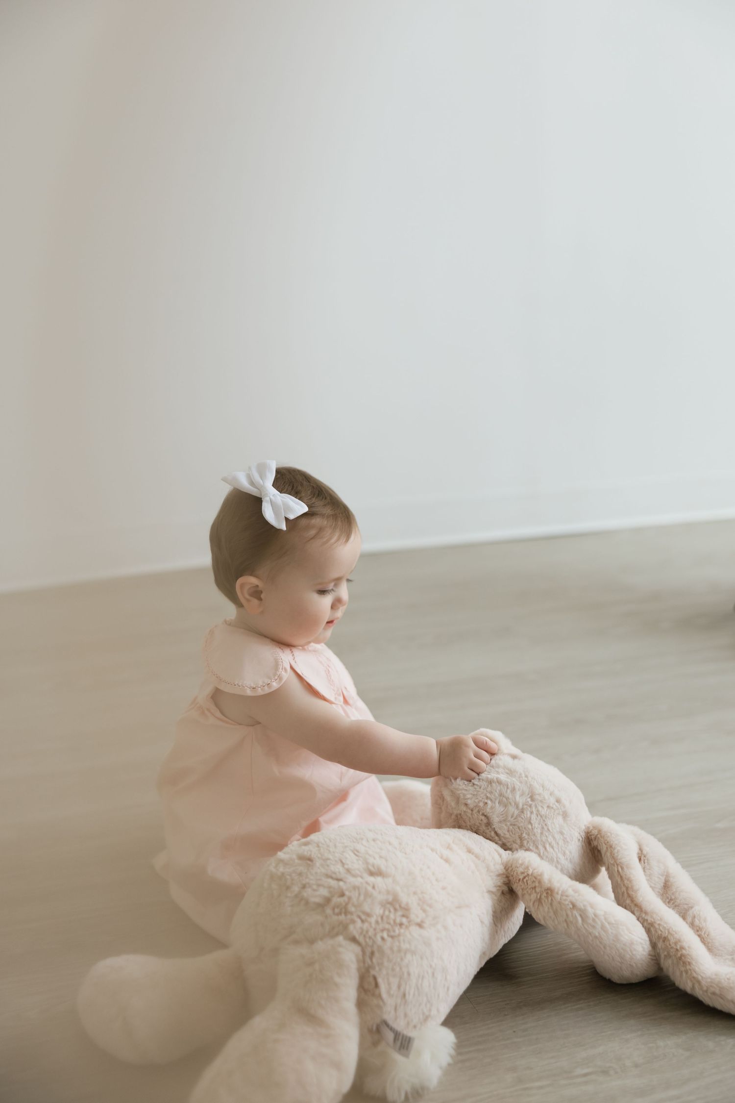 Baby girl in pink dress with white bow plays with large stuffed bunny on floor.