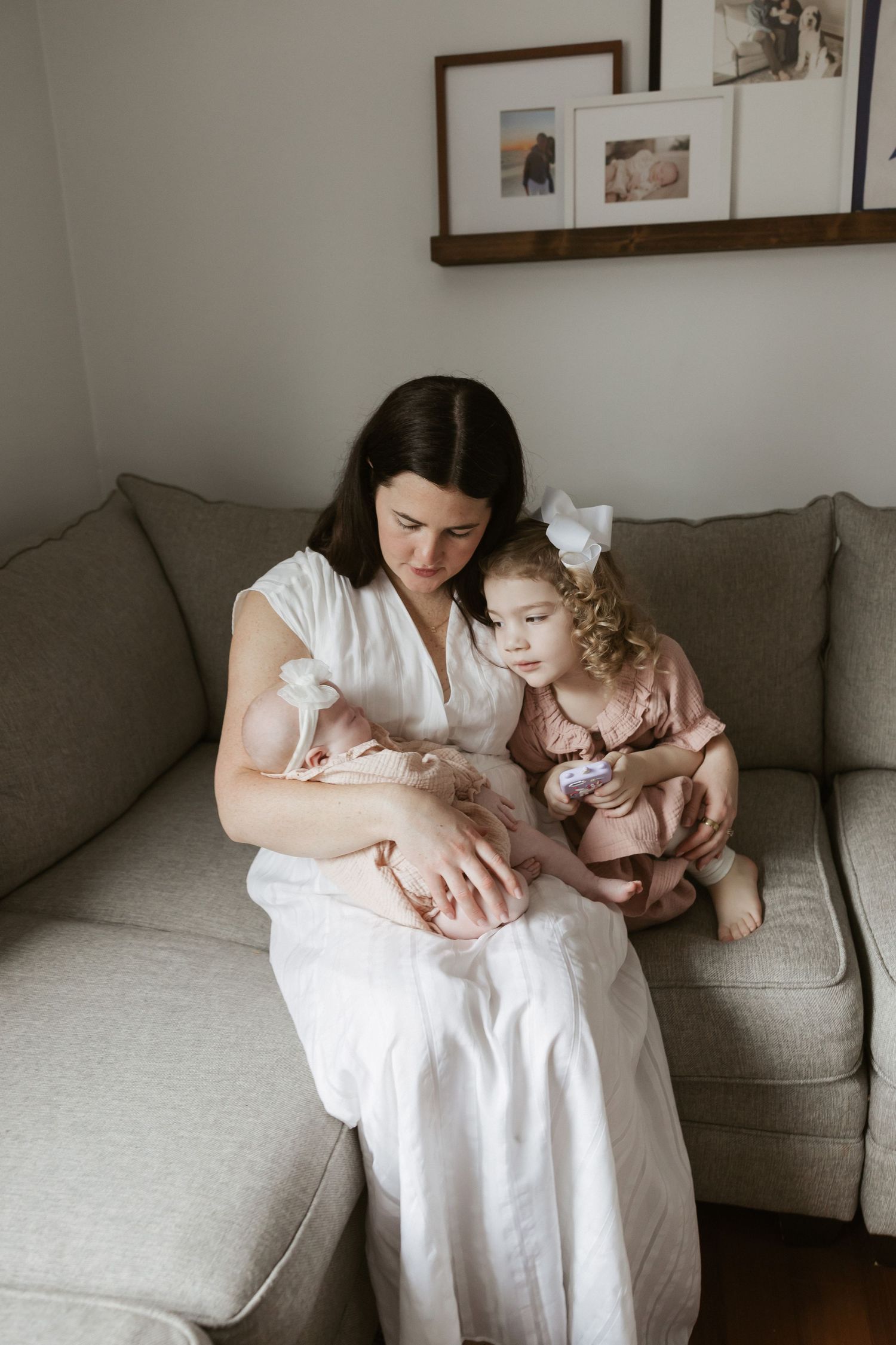 A mother in a white dress sits on a gray sofa holding a newborn while her toddler daughter in pink leans close.