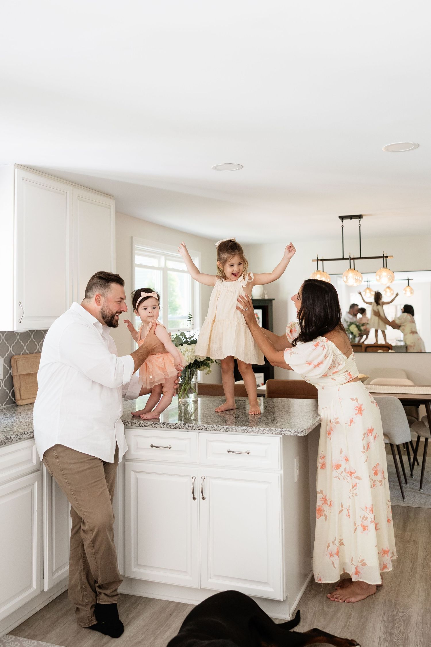 Family of four enjoying playful moment in modern white kitchen, toddler standing on island, parents smiling.