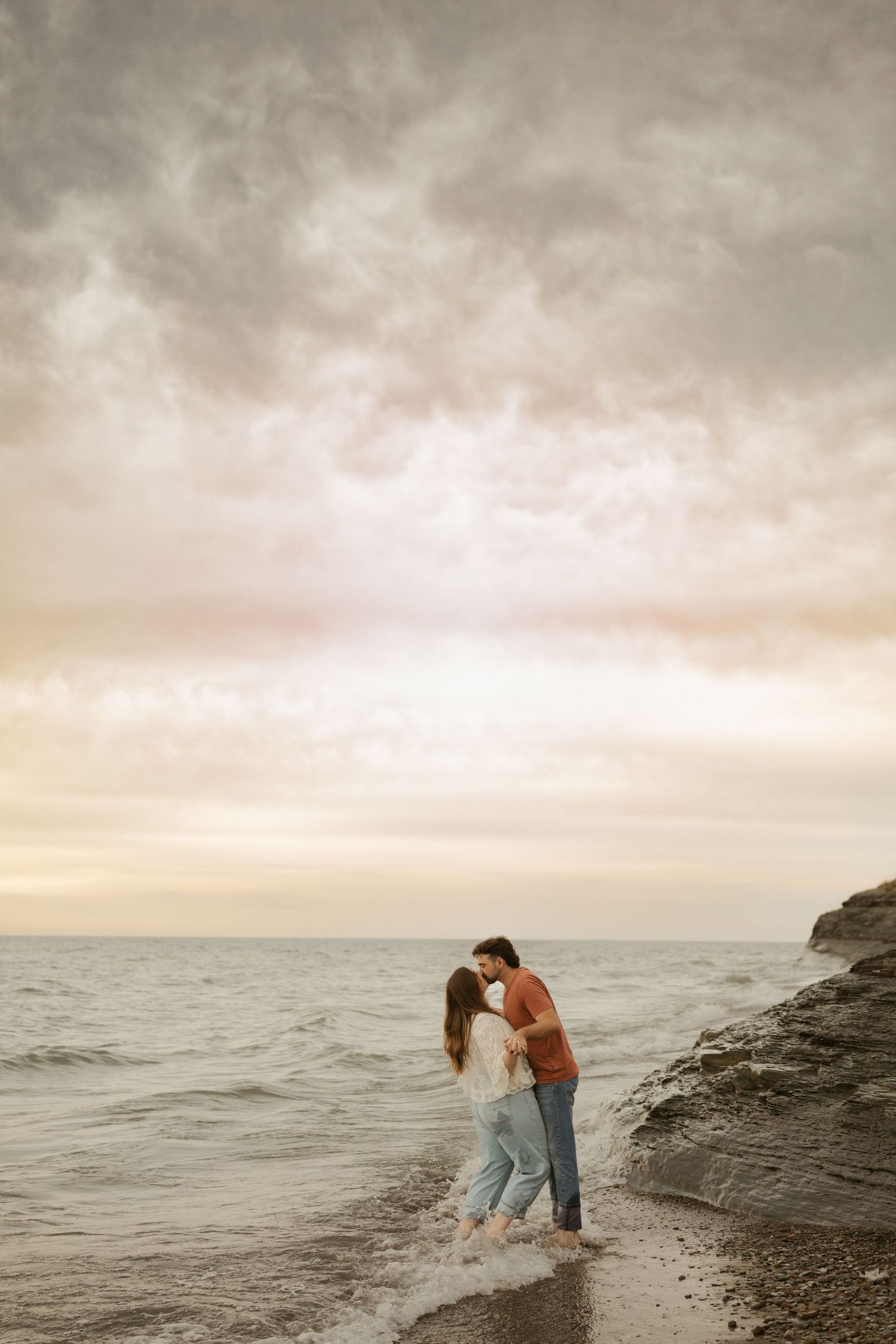 Romantic couple kissing at the ocean's edge during a dramatic sunset with moody clouds overhead.
