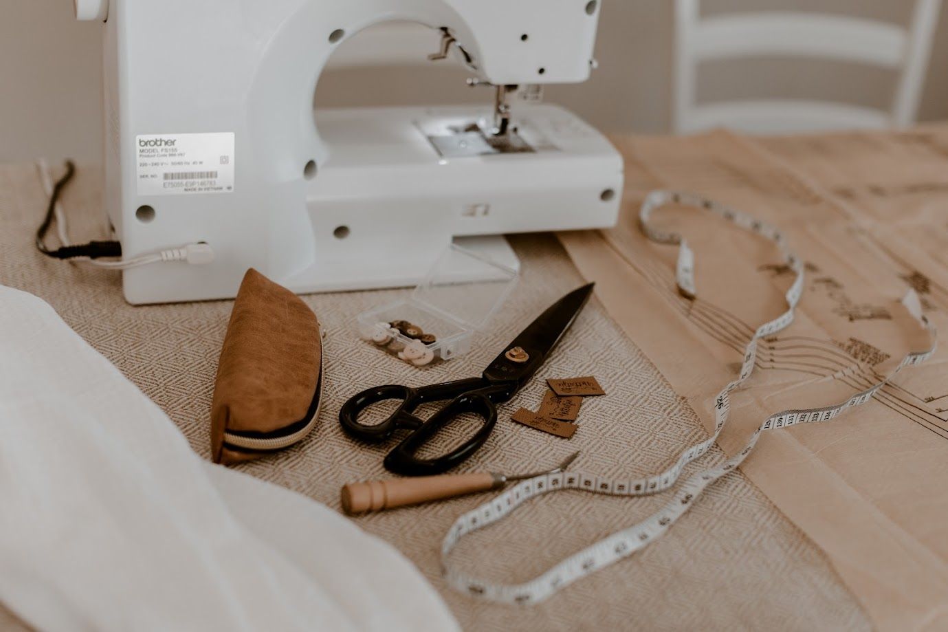Sewing machine, black scissors, measuring tape, and leather tools arranged on a burlap fabric surface for tailoring.