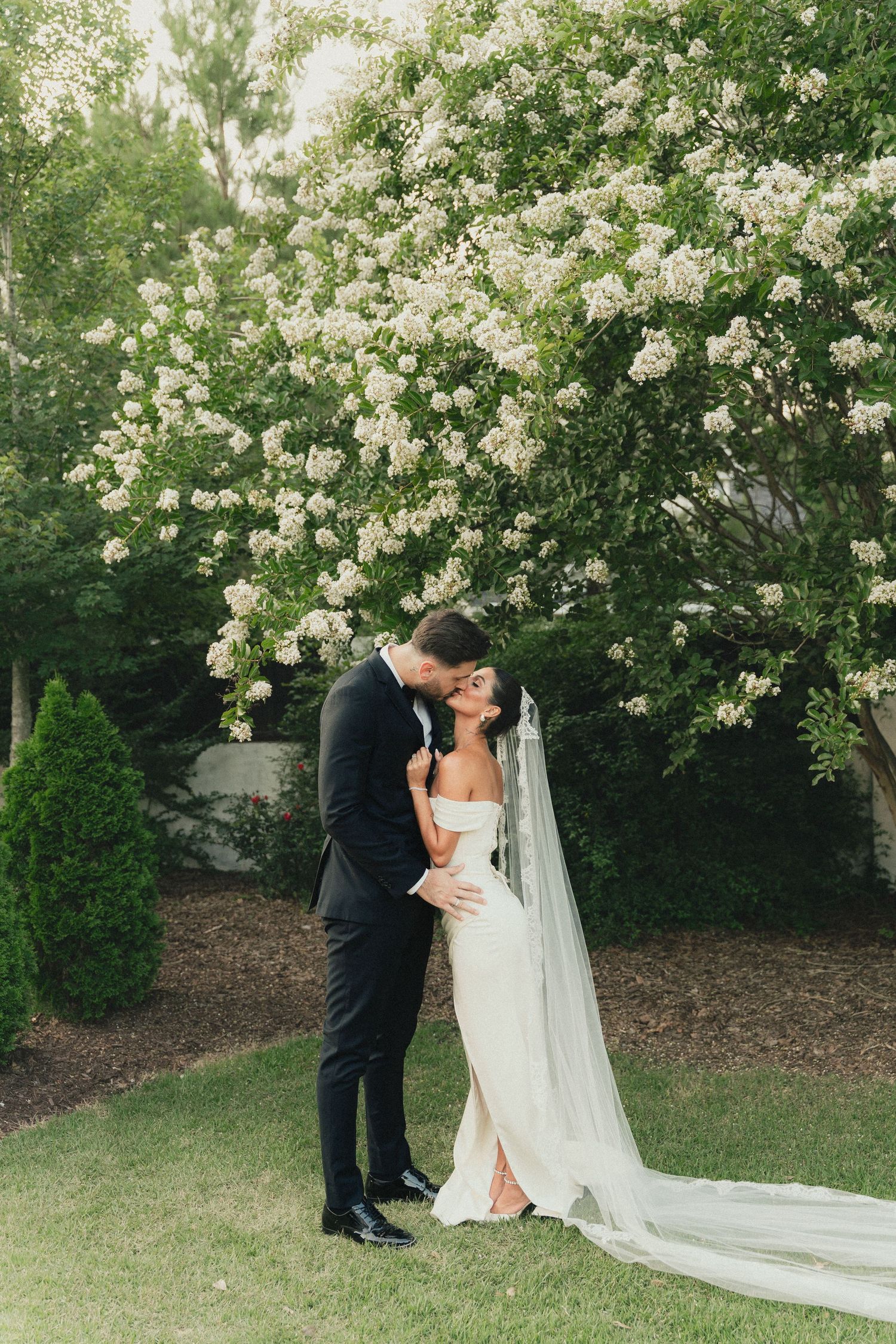 A romantic wedding couple shares intimate moments under blooming white flowers in a lush garden setting.