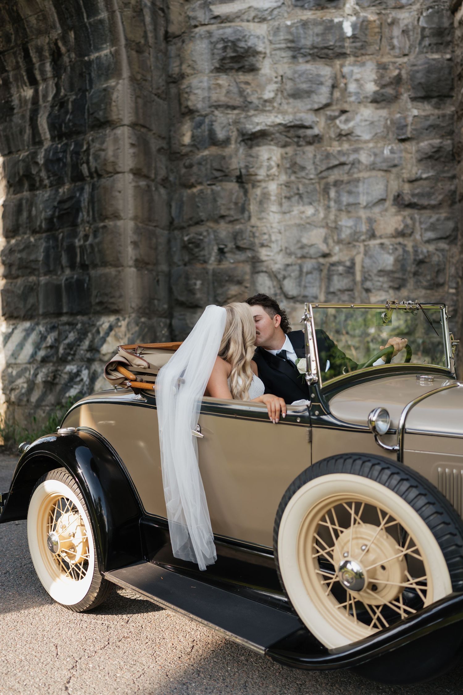 A vintage cream-colored classic car parked against a stone wall backdrop.